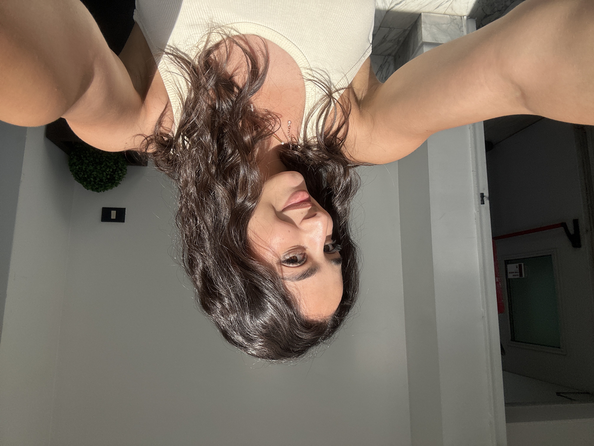 A medium shot selfie shows a woman with long, dark, wavy hair and tan skin, wearing a white tank top. The lighting is bright and sunny. Behind her is a white wall and a doorway leading to a darker area. To the upper left, a green, rounded artificial plant is visible, and a small black rectangle is attached to the wall below it. The location is an indoor space in Milan, Italy.