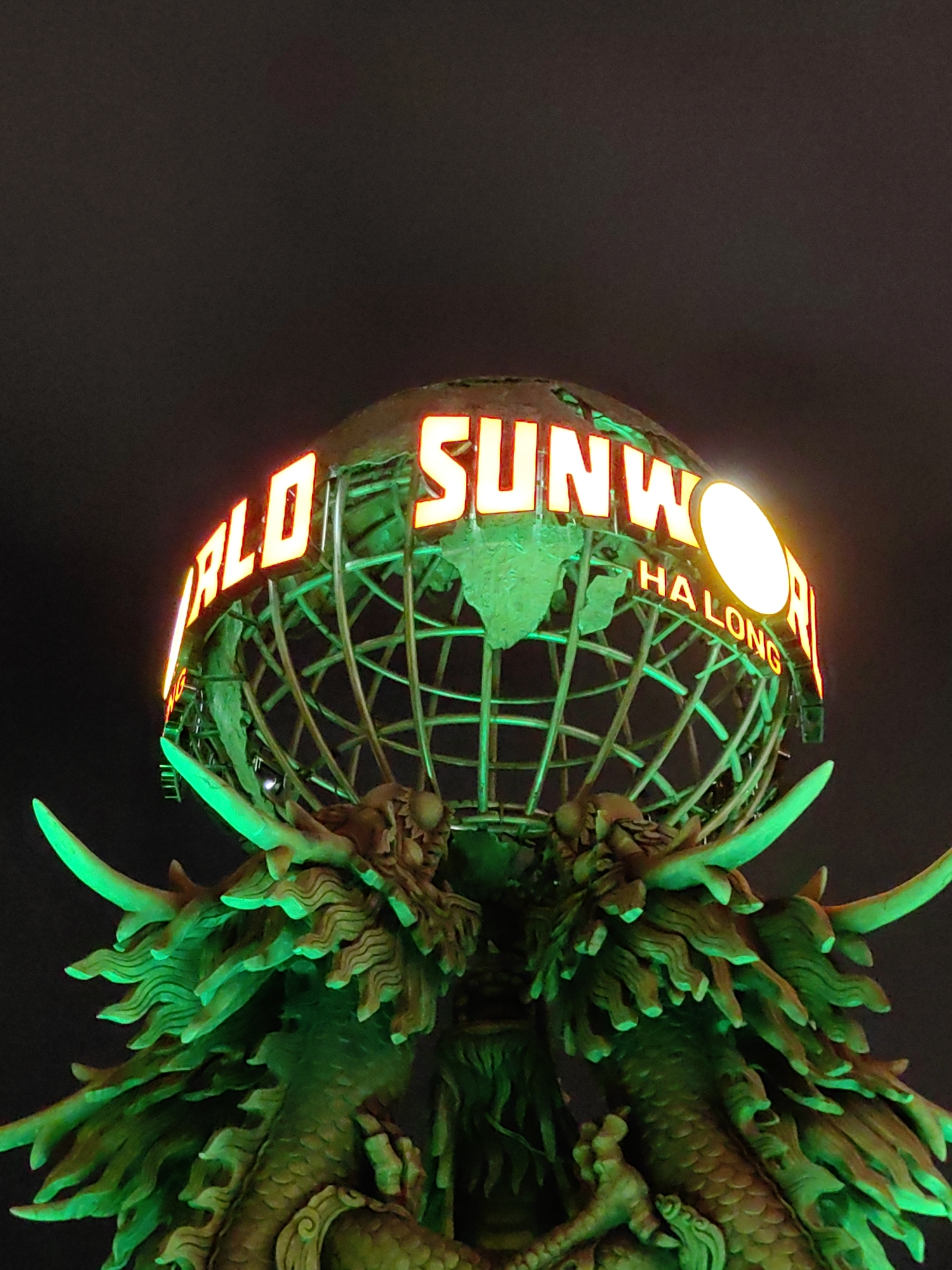 The image shows the "World Sunworld Halong" sculpture in Bai Chay Ward, Vietnam, at night. The structure features a metal globe with neon-lit letters spelling out the name. A pair of green dragon sculptures support the globe. The background is a dark, unlit sky.