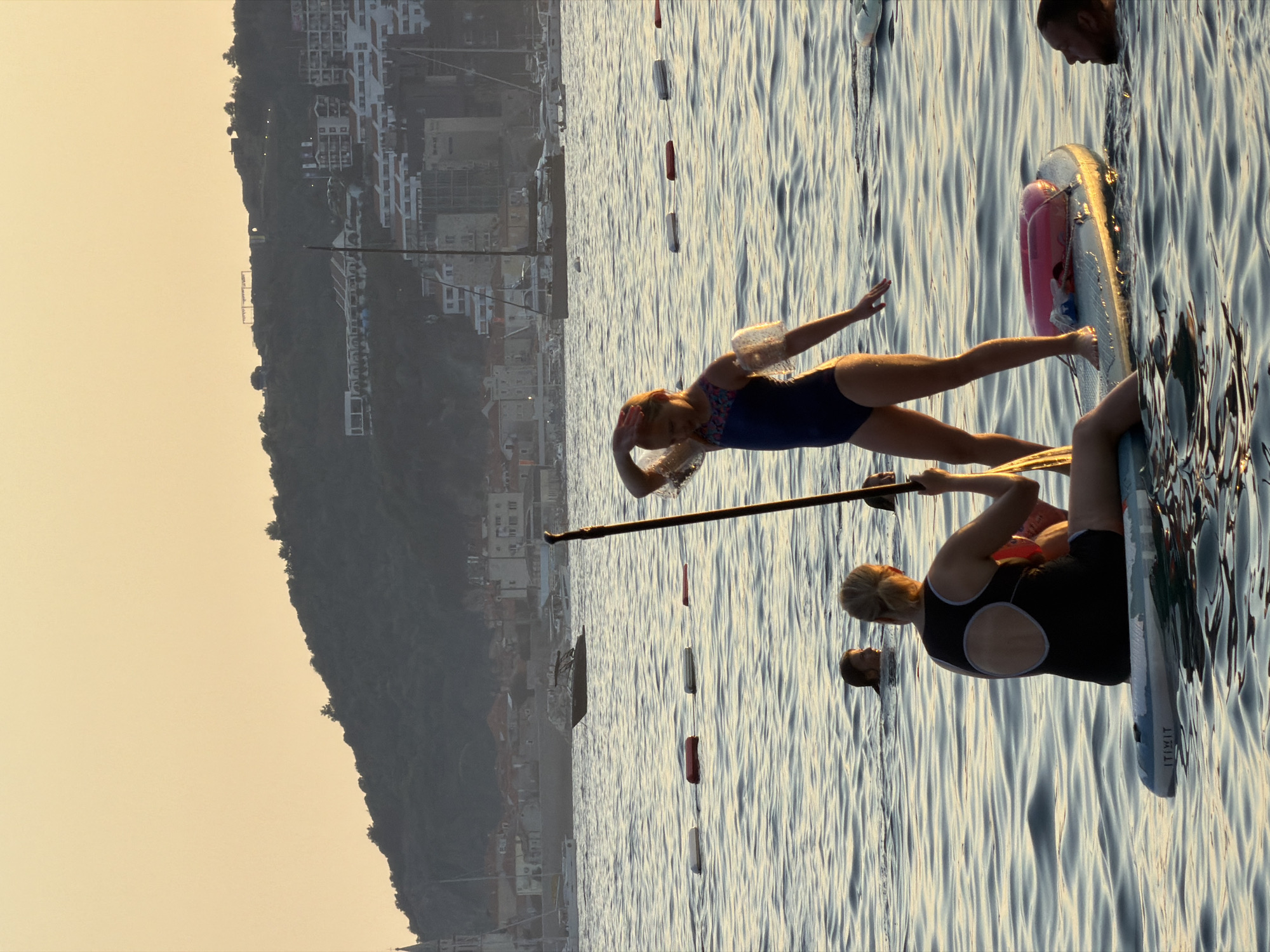 In the waters off Bečići, Montenegro, a group of people engage in water activities at sunset. In the foreground, a woman sits on a paddleboard, holding a paddle and looking towards a young girl standing on the same board. The girl is wearing a blue swimsuit and arm floaties. Another paddleboard is visible with a pink inflatable ring on it. A man's head emerges from the water to the right of this paddleboard. Further back, two individuals are swimming, their heads visible above the water. In the mid-ground, a line of buoys floats on the surface. The background features a hilly coastline dotted with buildings and trees, with several boats anchored in the bay. The sky is a warm, soft orange, characteristic of sunset.