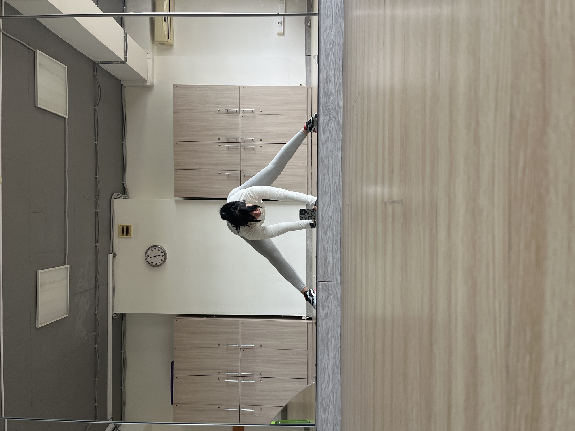 A woman is photographed in a mirror. The photo is taken from a high-angle perspective with a focus on the woman and her reflection.
The woman is wearing gray leggings, a white long-sleeved shirt, and black and gray sneakers. She is bending over, stretching her legs.
Behind the woman is a white wall with gray cabinets. On the wall to the left of the cabinets, there is a gray wall with a light fixture and visible wires. A clock is visible on the wall.
To the right of the woman is a wood-paneled wall. A horizontal metal bar is near the top of the frame. The location of the photo is not specified. Sochi, Russia is not explicitly specified, either.