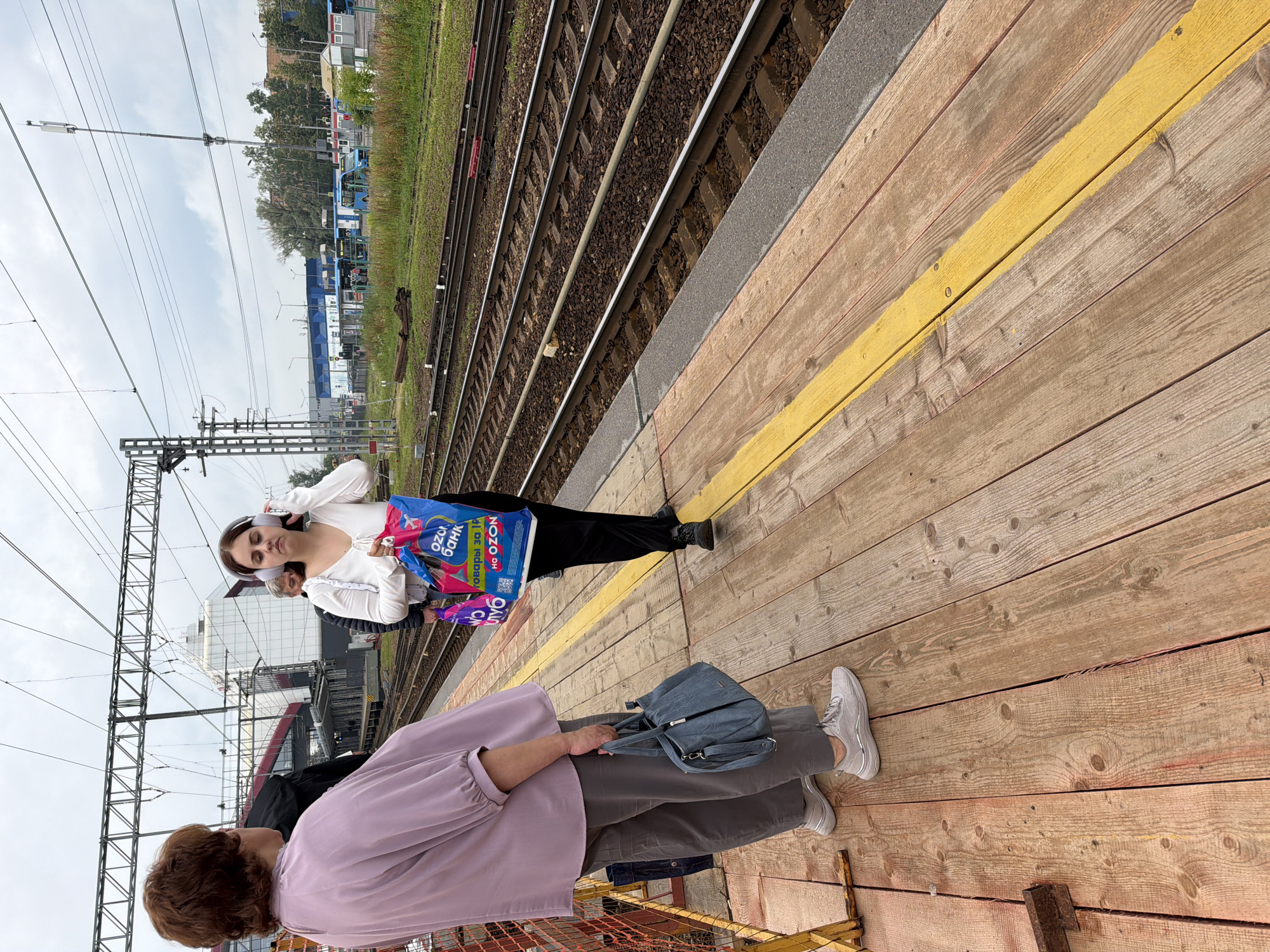 The image depicts a train platform in Shcherbinka, Russia, on an overcast day. Two women are standing on the wooden platform. The woman on the left, facing away from the camera, wears a light purple blouse and grey pants, carrying a blue handbag. The woman on the right, facing the camera, has on a white top and black pants, holding two colorful shopping bags. She is wearing headphones. The platform has a yellow line marking the safety edge. Train tracks run alongside the platform, with overhead electrical lines and a modern station building in the background. Buses and other vehicles are visible further in the distance, along with trees and industrial-looking structures. The overall scene suggests a typical day at a suburban train station.
