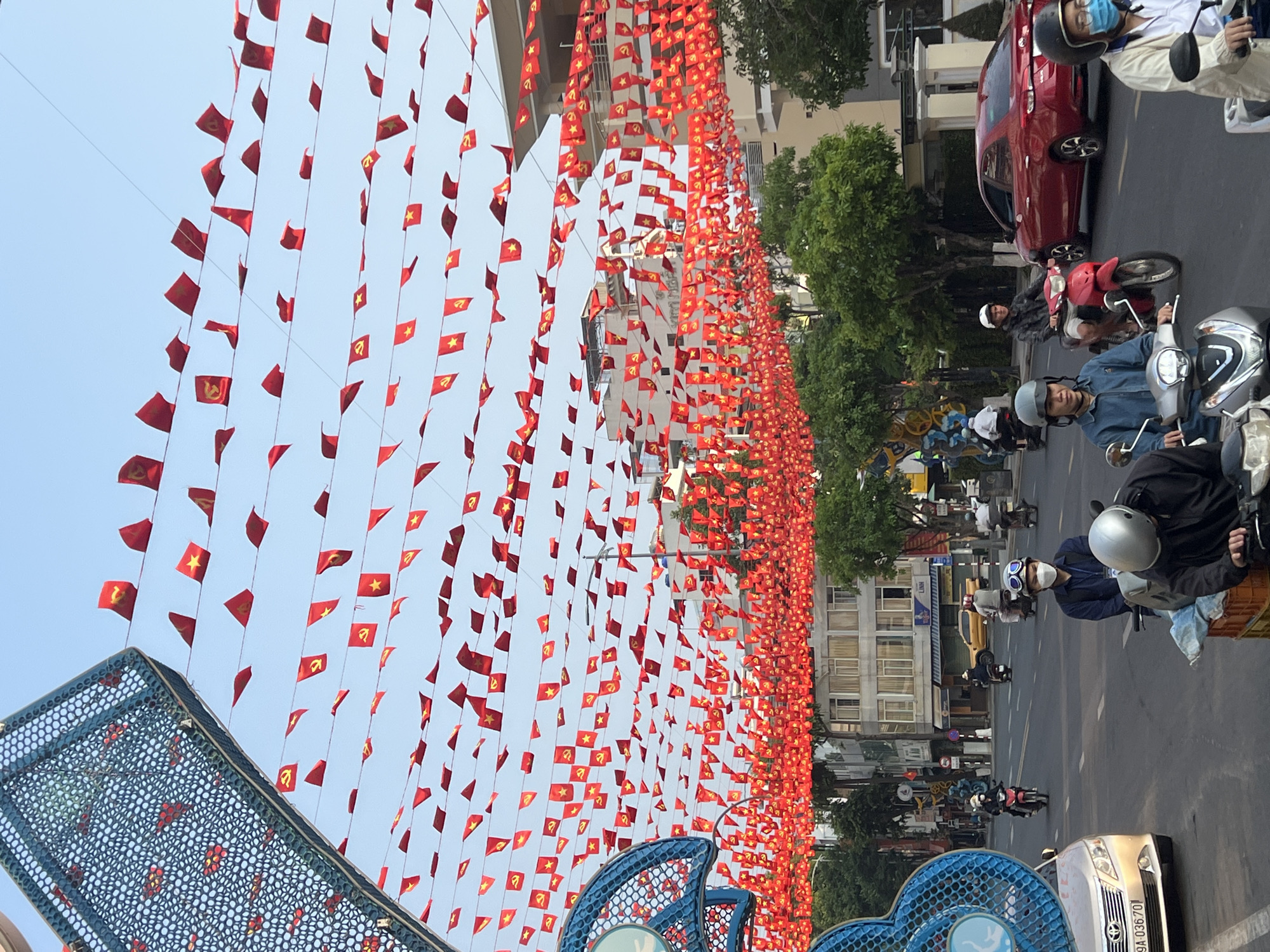The image captures a street in Nha Trang, Vietnam. Red flags, some bearing the Communist Party symbol and others a single gold star, are strung across the street, filling the sky above. The street is active with traffic, mostly motorbikes with riders wearing helmets. Buildings line the street, and trees provide some greenery. A red car is parked on the right side of the street. A blue, decorative structure is visible in the foreground. The sky is light blue, suggesting it is daytime. The overall scene presents a bustling urban environment decorated for a patriotic occasion.