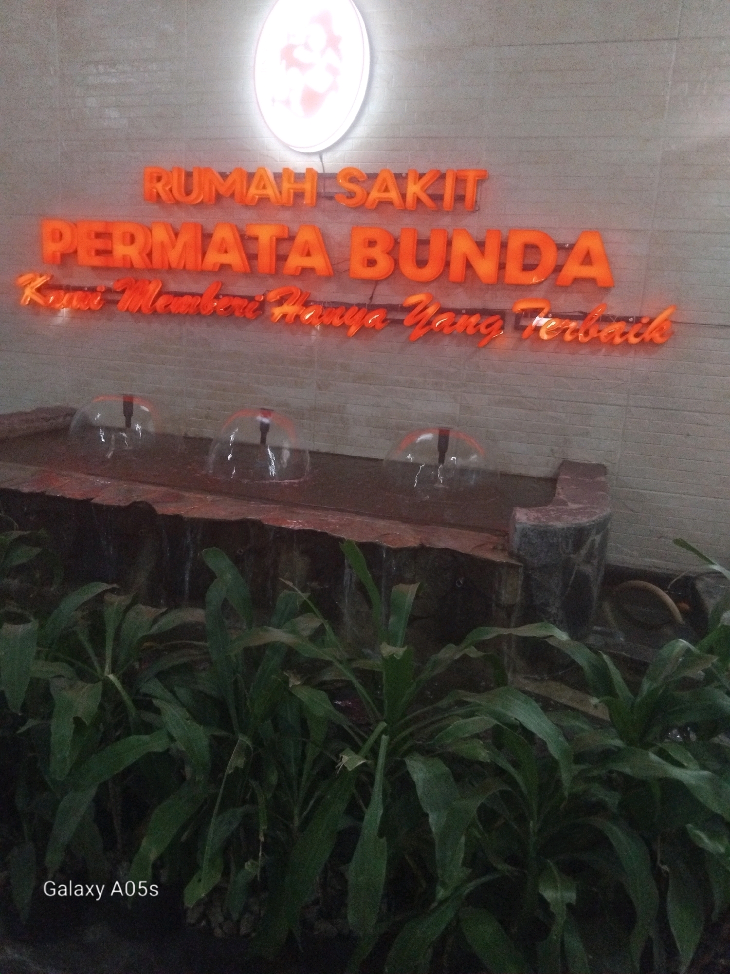 In the city of Purwodadi, Indonesia, a neon sign reads "RUMAH SAKIT PERMATA BUNDA" (Permata Bunda Hospital). Below this, another neon sign states, "Kami Memberi Hanya Yang Terbaik" (We Give Only The Best). In front of the sign, there is a small water fountain with three bubbling water features. Behind the fountain, lush green plants are visible. The background wall is made of textured bricks. The image is branded with "Galaxy A05s" at the bottom left.