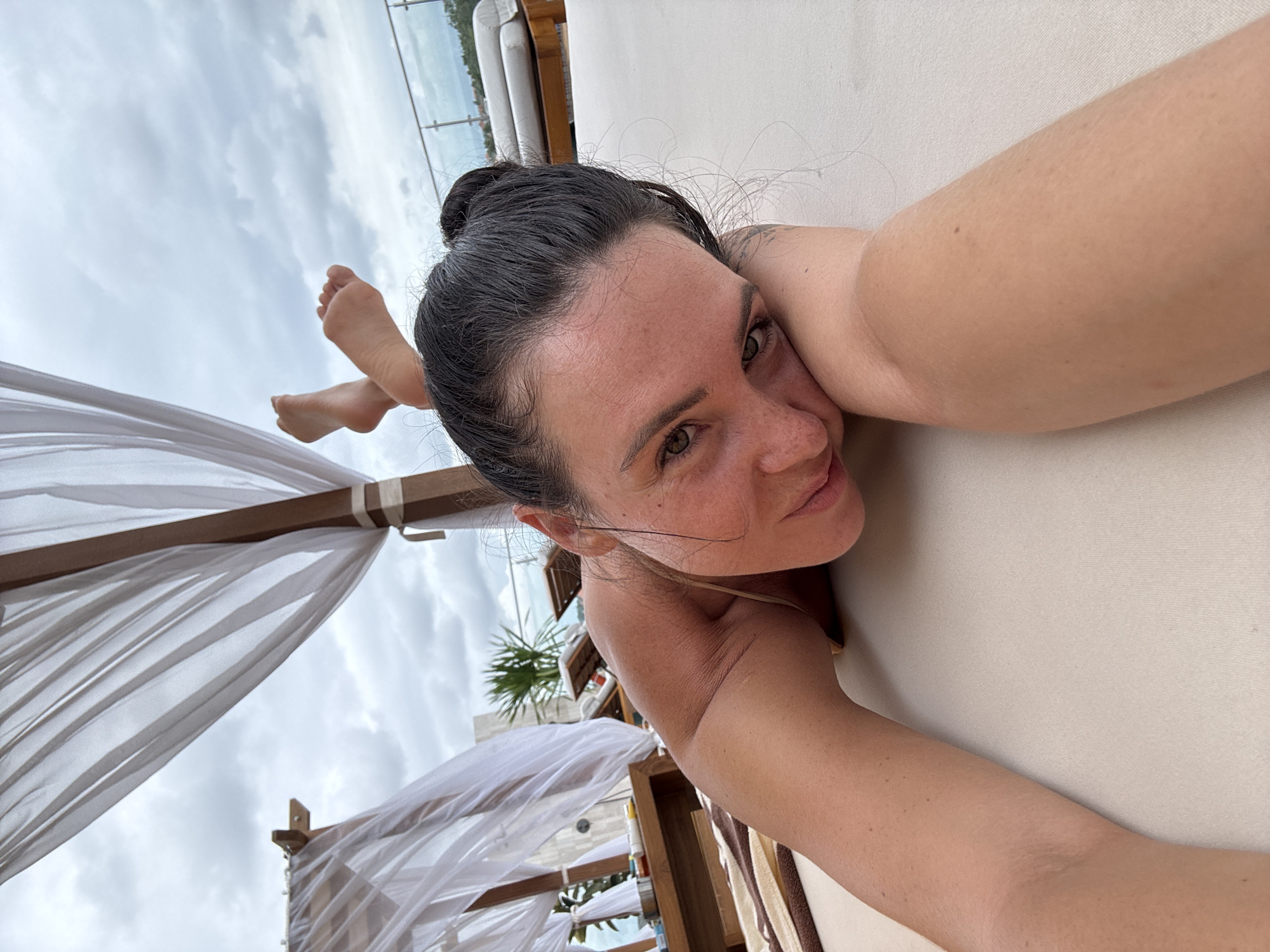 A woman lies prone on a beige lounge chair outdoors, with her head resting on her arm. Her dark hair is pulled into a bun. Her feet are crossed and visible in the upper center of the frame against a cloudy sky. She wears a bikini top. The setting appears to be a rooftop lounge area, with white sheer curtains draped from a wooden structure in the foreground, and lounge chairs and railings visible in the background. There are some small bottles and containers on a wooden shelf to the left. The scene suggests a vacation or resort environment.