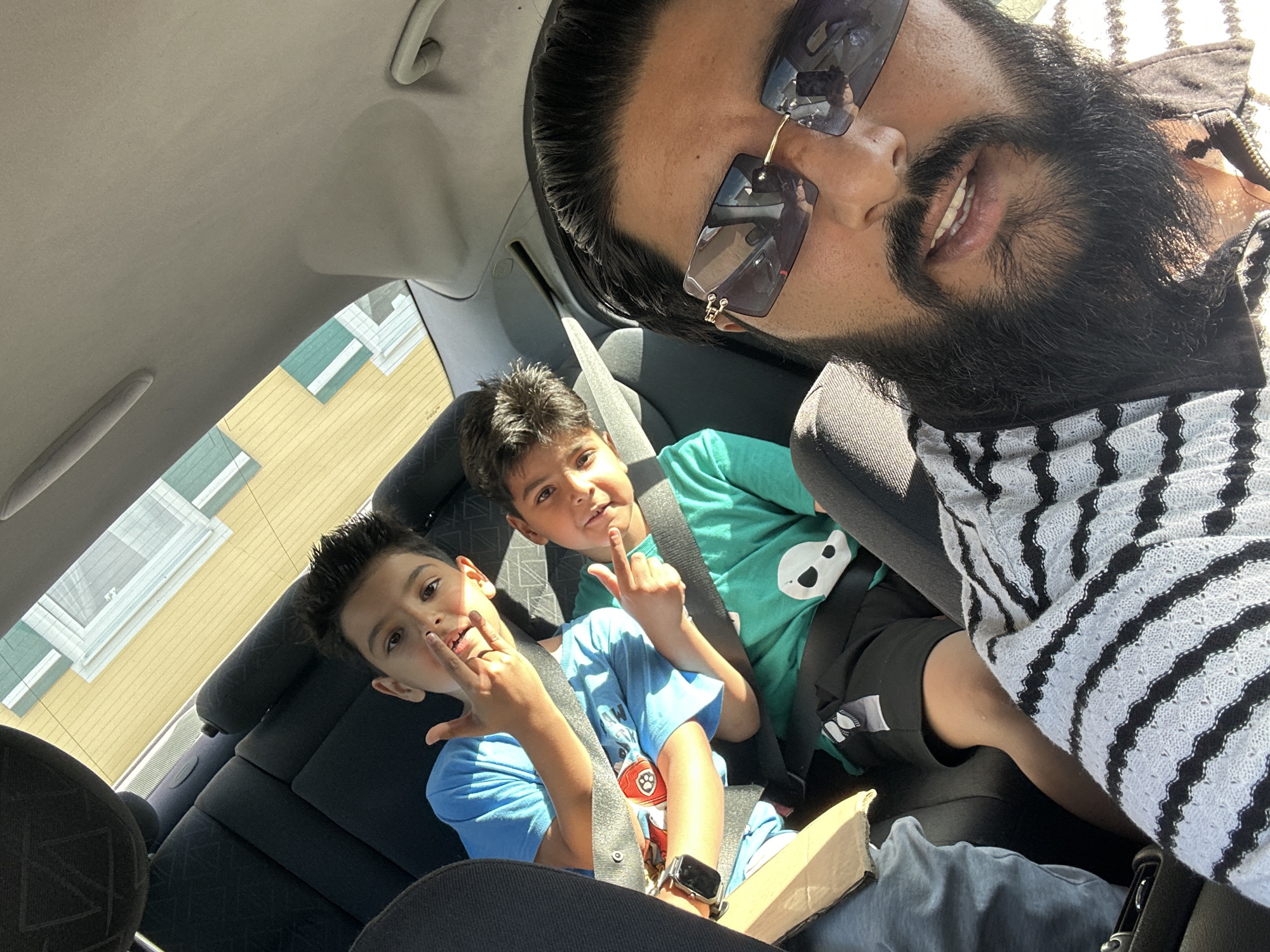 A selfie is taken inside a car. In the foreground, a man with a beard and sunglasses is visible. Behind him, two young boys are seated in the back seat, both wearing seatbelts. One boy is wearing a blue t-shirt, and the other is wearing a green t-shirt. The car's interior is visible, including the back seats and part of the dashboard. Outside the car windows, a building with yellow and green sections can be seen, suggesting an urban or suburban environment. The image is well-lit, indicating it was taken during the daytime. There is no indication of the location being Mainburg, Germany.