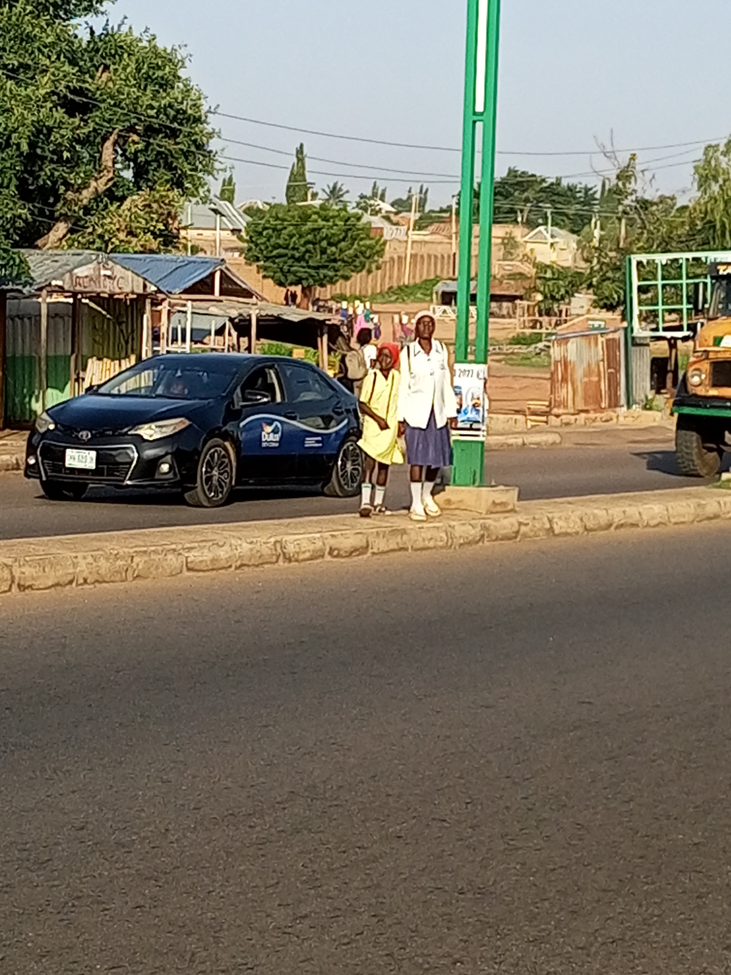 In Garko, Nigeria, two young individuals, one in a yellow dress and the other in a white shirt and blue skirt, stand on a paved sidewalk next to a black Toyota sedan. The car, bearing the license plate "HK 633 K," has a blue graphic on its side. Behind them, a green pole with a poster attached is visible. Further in the background, a yellow and green truck is parked. The scene unfolds near a street with buildings and trees lining the horizon under a clear sky.