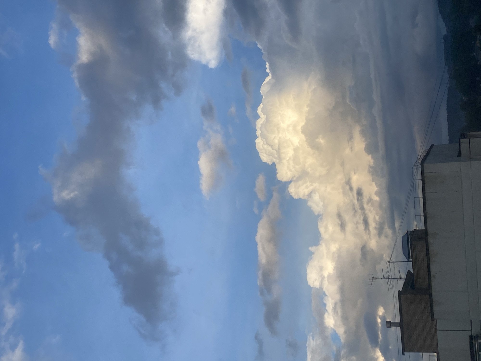 The image displays a dynamic sky scene over an urban area, rotated counter-clockwise. The dominant feature is a vast expanse of sky filled with a mix of large, dramatic clouds and clear blue patches. Prominent bright white clouds, likely cumulus, are strikingly illuminated by what appears to be late afternoon or early evening sunlight, creating deep shadows and textural contours. Interspersed are darker grey clouds, suggesting varying cloud densities or areas obscured from direct sunlight.
In the lower right section of the frame, a multi-story building is visible, featuring a flat roof with brick structures, a small blue utility box or tank, and several antennas or satellite dishes. Thin utility lines stretch diagonally across the upper right, connecting to the building. In the far distance on the right, a dark, forested mass resembling hills or mountains is silhouetted against the sky. The lighting suggests a time of day with strong, directional sunlight, indicating either sunrise or sunset, likely the latter given the warmth of the light on the clouds. The weather appears active and visually dramatic due to the varied cloud formations. No visible text is present in the image. The location context for this scene is Chaikovsky, Russia.