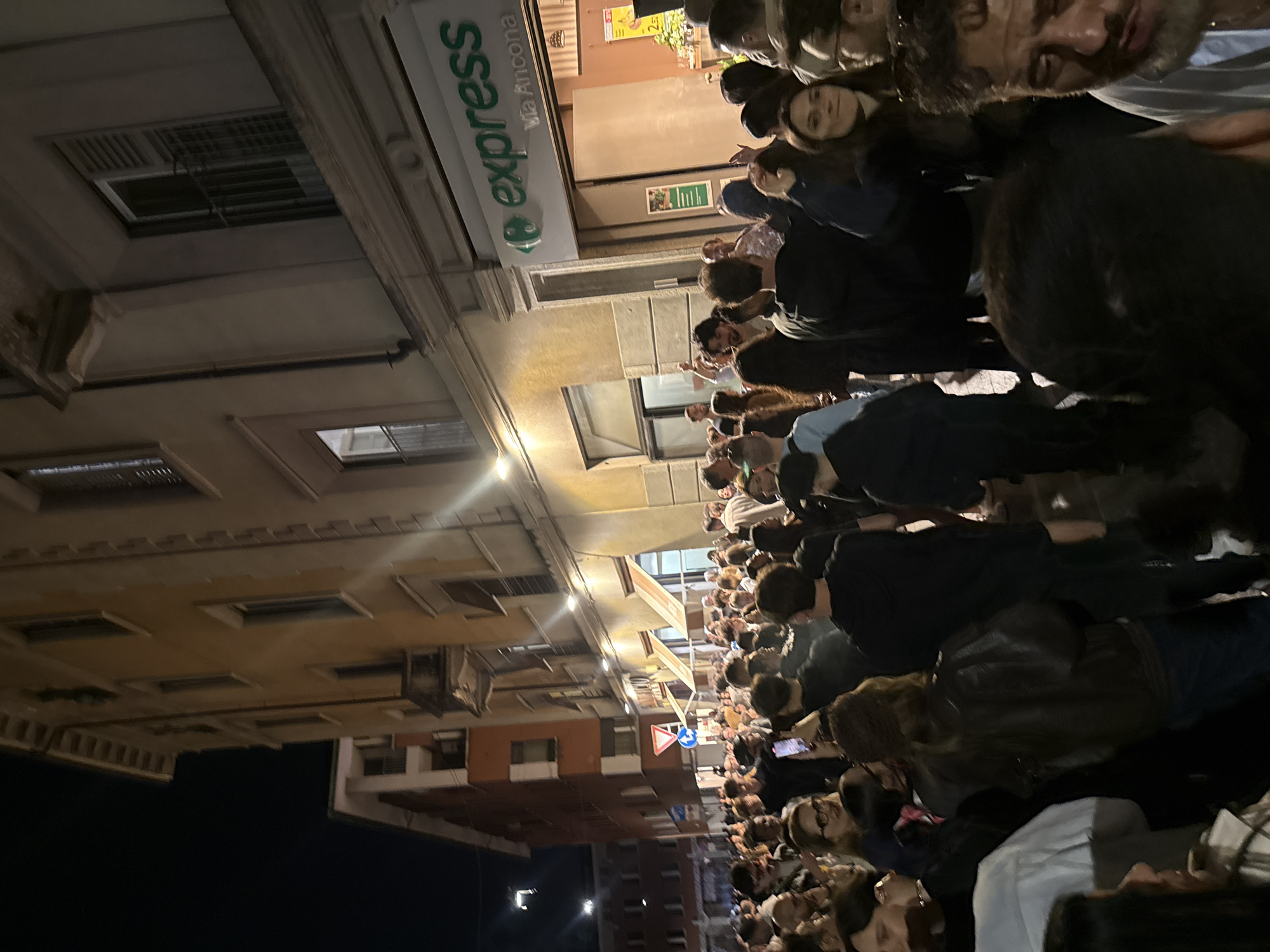 This media file depicts a vibrant night scene on a busy street, likely in Milan, Italy. The image is rotated 90 degrees counter-clockwise.
The primary subjects are a large, dense crowd of people, mostly young adults, filling the street from the foreground into the midground. They appear engaged in social activity, standing and conversing, suggesting a lively gathering or a typical evening street scene. Their faces are illuminated by artificial lights, and their attire is casual.
The setting is an urban street flanked by multi-story buildings. On what would be the upper left side of an upright image, a prominent "Carrefour express" store sign is clearly visible. Below the store name, the text "Via Ancona" is displayed, directly confirming the location in Milan, Italy. Near the store entrance, a small sign shows "2€." Other buildings feature multiple windows, with one building having a light, possibly yellow, facade and another in the distance appearing reddish-brown. Streetlights illuminate the scene, casting glows on the buildings and the crowd against a dark night sky. A blue circular street sign with a white arrow is visible further down the street.
The time of day is clearly night, evidenced by the darkness and the reliance on artificial lighting. The mood appears relaxed and social, without any indication of specific events like protests or performances, but rather a bustling street atmosphere.