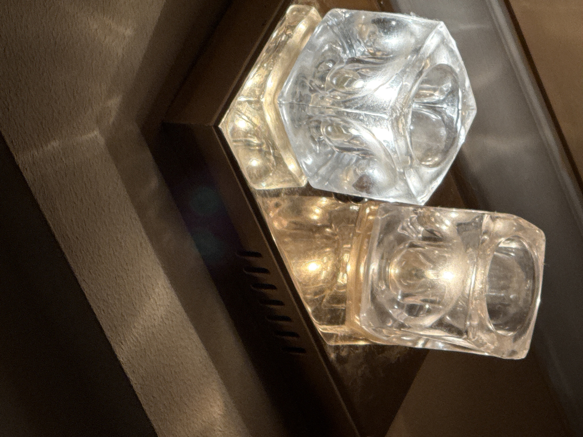 The image shows a close-up view of two glass light fixtures mounted on a ceiling. The fixtures are cube-shaped with faceted glass. One fixture appears to be lit, emitting a warm yellow light, while the other is unlit and reflects the surrounding environment. The fixtures are mounted on a metallic base, possibly brass or gold-colored, which is attached to a darker, textured ceiling. The lighting is dim, creating shadows and highlights on the glass. There are no people or animals visible in the image. The location of the image is not specified in the original prompt and cannot be determined from the visual information provided.