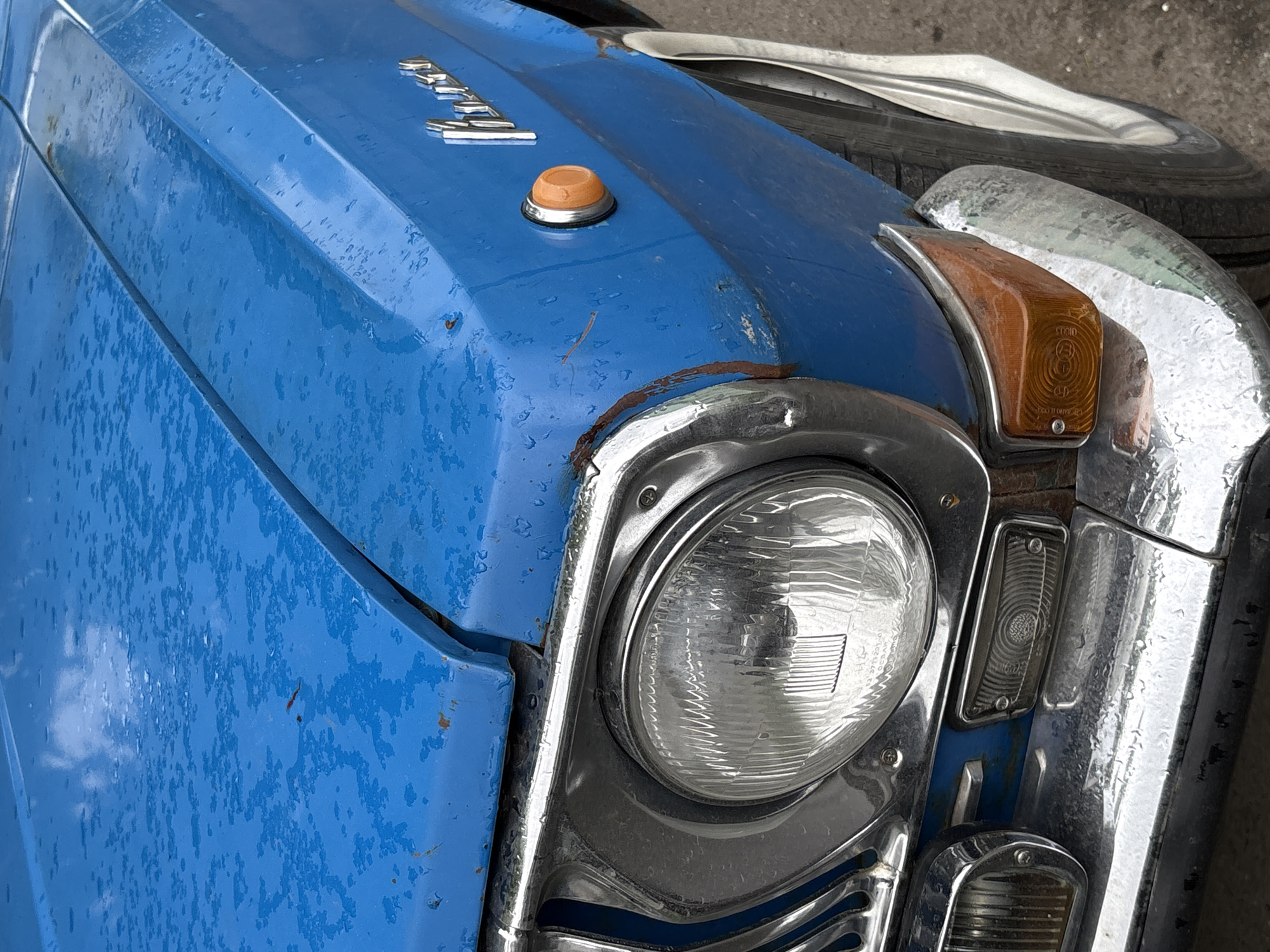 The image shows the front left corner of a vintage, light blue car. The car is heavily weathered with chipped paint and rust present around the headlight and hood. The headlight is round with a chrome surround. An amber turn signal is positioned next to the headlight. The car's hood has a raised section and the word "Moskvich" is spelled out in chrome lettering. A small amber light or button is visible on the hood. The surrounding environment is unclear, with only a tire partially visible in the upper right corner. There is no indication of the location being Saint Petersburg, Russia.