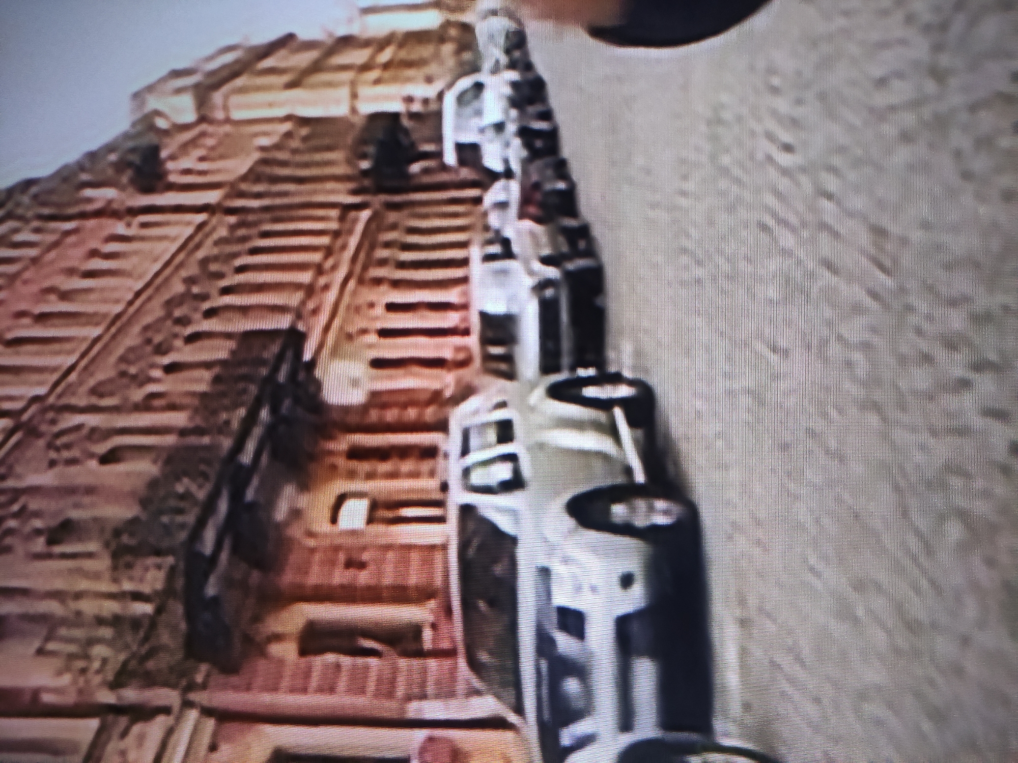The rotated image depicts a line of parked vehicles alongside a massive, multi-tiered structure in Pavlodar, Kazakhstan, under a clear daytime sky. The structure, visible on the left, appears to be a large industrial building or complex, characterized by numerous horizontal elements suggesting multiple floors or ledges, and a weathered, reddish-brown facade.
A row of approximately five to seven light-colored vehicles, resembling white or silver vans or SUVs, are parked in sequence adjacent to the building. One vehicle in the foreground shows discernible text on its side, which appears to read "POLICE" or a similar variation, along with additional characters such as "0J0." This suggests the vehicles may belong to law enforcement or security services.
A blurred hand or arm is partially visible in the upper right foreground, indicating a close perspective, possibly from a person holding a camera. To the right of the vehicles, a textured, lighter-colored surface is visible, likely a wall or ground. The scene is static, with no discernible activity or people in motion, primarily capturing the parked vehicles and the imposing architecture of the background structure.