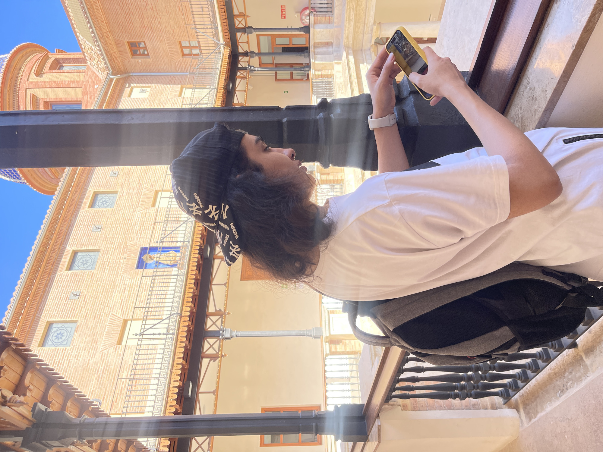 The image depicts a person holding a yellow smartphone, appearing to be taking a picture or recording a video. The person is wearing a white t-shirt and a black and white baseball cap. A gray and black backpack rests behind them, against what appears to be a stone railing. The scene is set in València, Spain. In the background are several multistory buildings with brick facades. One building has a dome with blue and white tiling. There are windows with intricate designs, wrought iron balconies, and wooden accents.