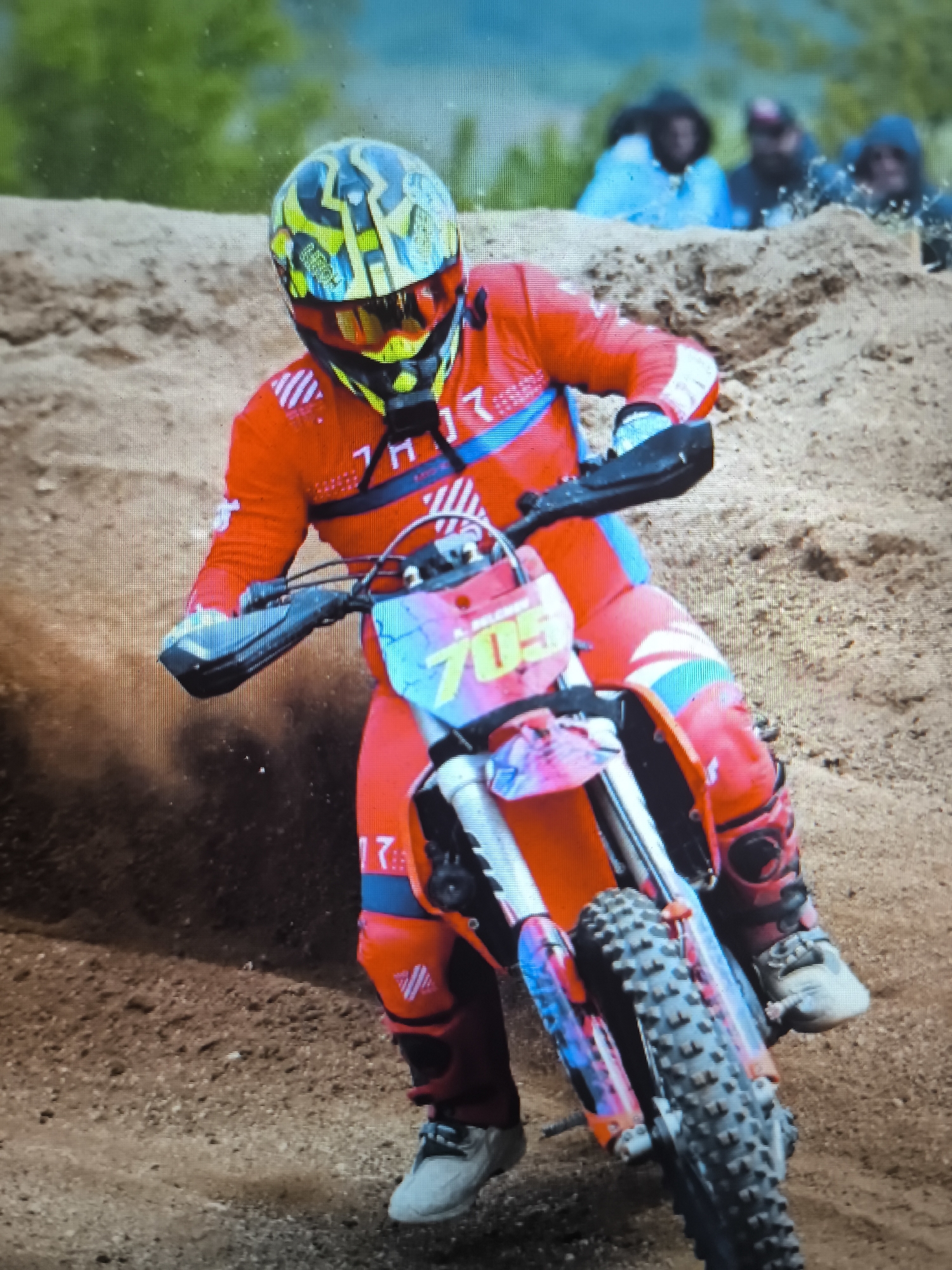 A person in an orange racing suit and helmet with blue accents is riding a dirt bike with the number "705" on a dirt track. The rider is leaning into a turn, kicking up dirt on the left side of the frame. In the background, a dirt berm is visible, behind which are several people. The photograph was probably taken in Enina, Bulgaria.