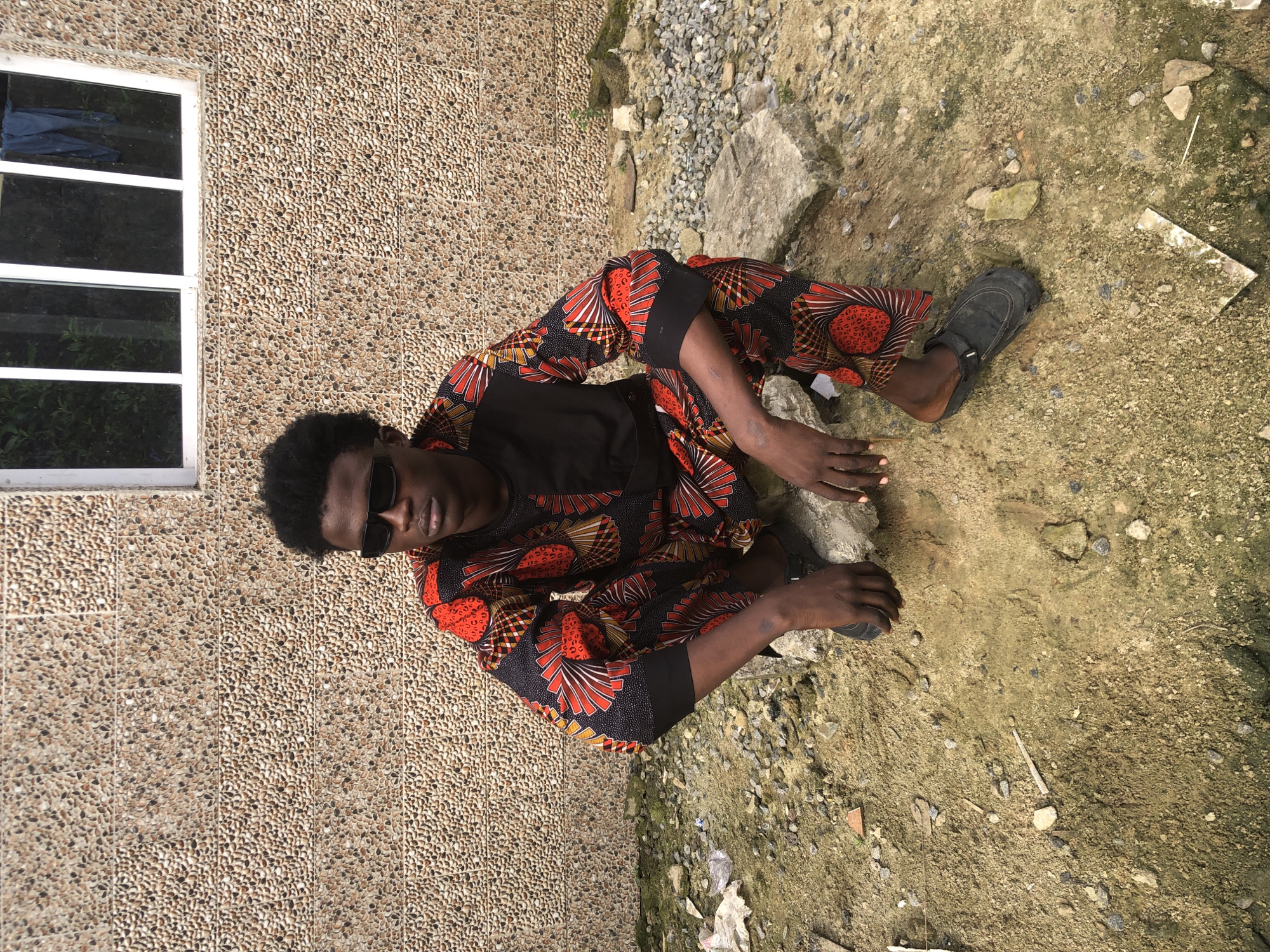 A young Black male, appearing to be in his late teens or early twenties, is crouched on a rocky, sandy patch of ground in front of a textured, light brown wall. He is wearing a black t-shirt underneath a vibrant, patterned garment with red, orange, and yellow designs, and black pants with a similar pattern. He has dark, curly hair and is wearing black sunglasses. His feet are clad in black slip-on shoes. The wall behind him has a window with multiple panes, and some dark blue fabric is hanging from the right side of the window frame. The ground is uneven, with scattered rocks, debris, and patches of dry grass. The overall impression is of an outdoor urban or semi-urban setting.