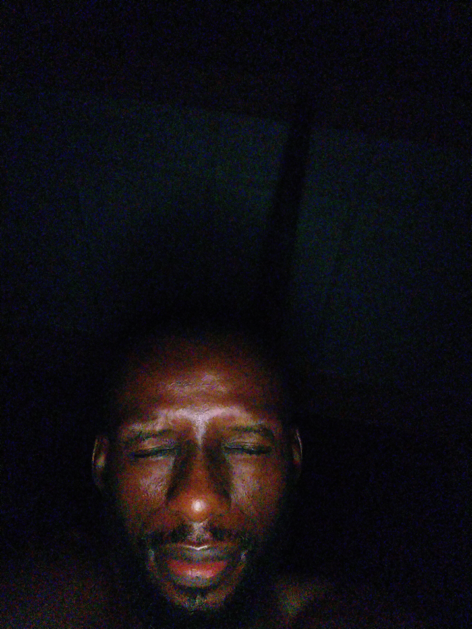 The image shows a close-up of a Black man's face with his eyes closed. The lighting is dim, with some warm light illuminating his forehead, nose, and lips. The background is dark and appears to be a plain surface. There is a faint vertical shadow on the right side of the background. The man has a short beard and mustache. His skin has a reddish-brown tone. The image is grainy, suggesting it was taken in low light conditions. The man's expression suggests he is relaxed or sleeping. There are no identifiable landmarks or features that would indicate the specific location within Uyo, Nigeria. The scene is indoors, and the environment is nondescript.