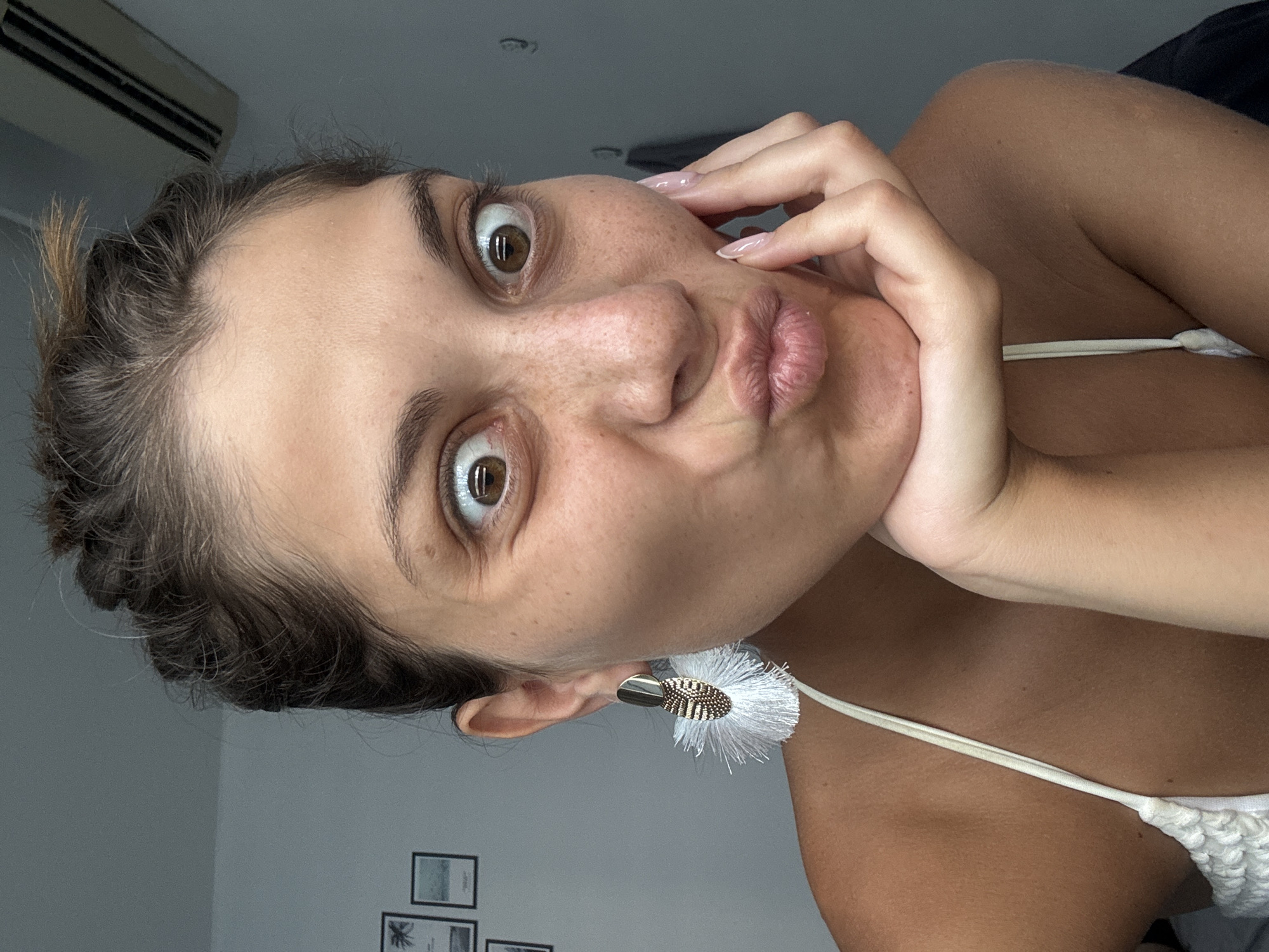 A woman with freckles is posing indoors. She has brown hair in braids and is wearing a white top and white earrings with tassels. She is making a duck face and holding her hand to her cheek. In the background, there is an air conditioner and a wall with pictures.