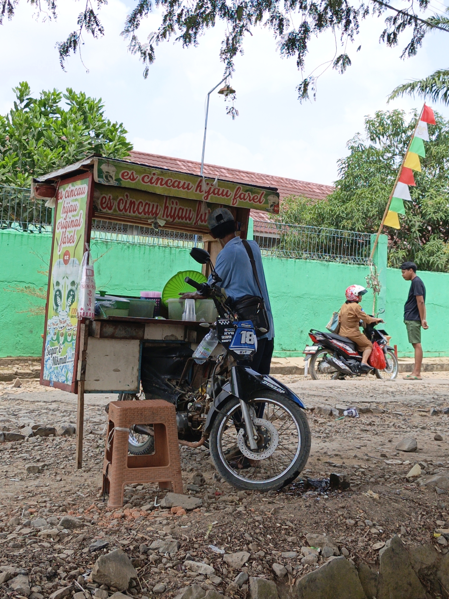 This image depicts a street scene in Gedung Nyapah, Indonesia, with a mobile beverage cart as the central subject. The cart, which appears to be a motorcycle with a custom-built structure attached, is parked on a gravelly street. It features signage for "es cincau hijau" (green cincau jelly ice). A man in a blue shirt stands behind the cart, attending to it. In the background, a woman in a brown uniform and helmet is seated on another motorcycle, facing away from the viewer. A man in a black t-shirt and shorts stands to the right of the woman, looking at her. A tri-colored flag is planted next to the woman's motorcycle. The environment is characterized by a green painted wall, trees, and a sky with scattered clouds. The ground is covered in stones and dirt.