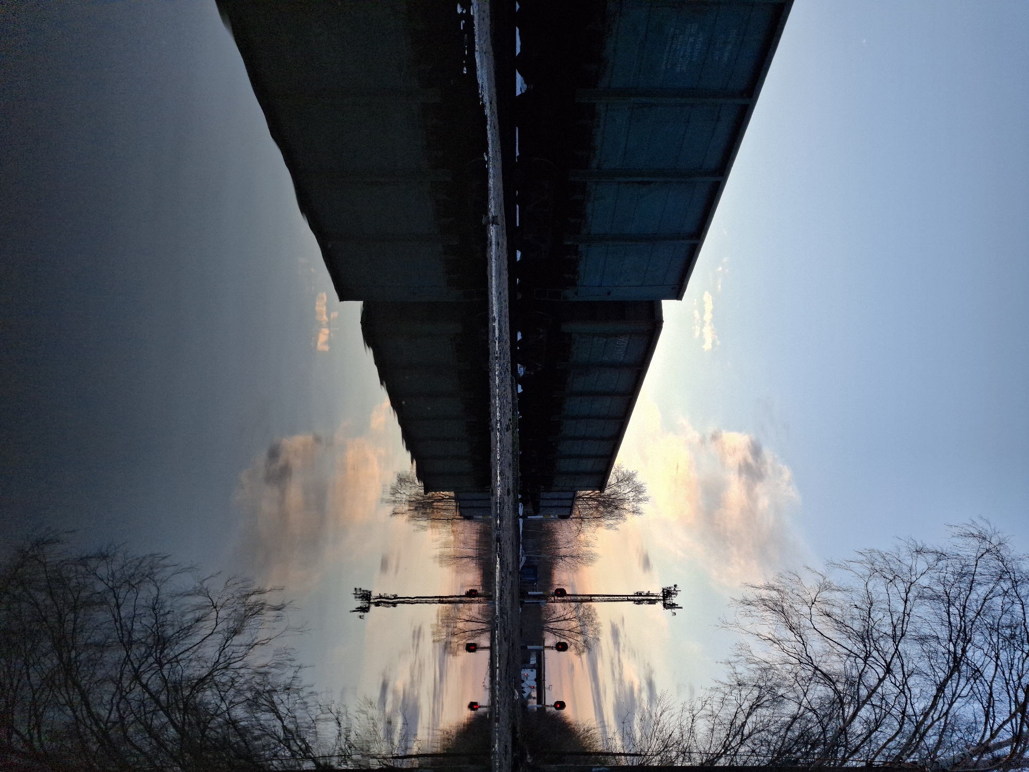The image depicts a reflection of a railway bridge and signal in water, likely a river or lake. The bridge is a dark, rectangular structure spanning the water, with train cars visible on top. The signal is a cross-shaped structure with red lights. Bare trees are visible on either side of the reflection. The sky is a gradient of blue and pink, suggesting dawn or dusk. The reflection creates a symmetrical effect, with the sky and trees appearing below the bridge and signal. Given the city of Chaikovsky, Russia, it is likely the Kama River or a major tributary.