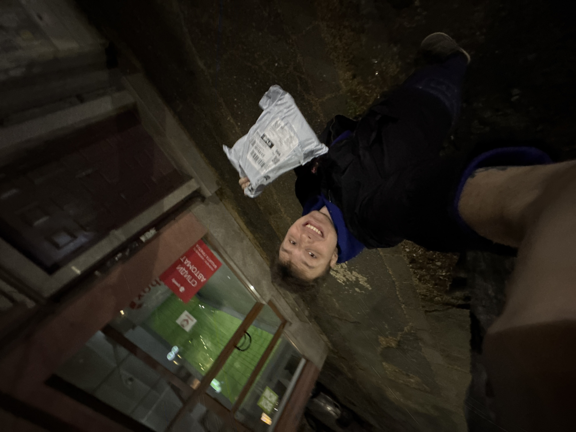 A person smiles while holding a package in their left hand. They stand outside of a store or business with a window display featuring green and white elements and a red sign with text in Cyrillic script. The location is likely within the city of Plovdiv, Bulgaria, based on the Cyrillic text on the signage. The photograph appears to be taken at night or in low-light conditions. The person wears dark clothing, including trousers and a jacket with a blue hood.