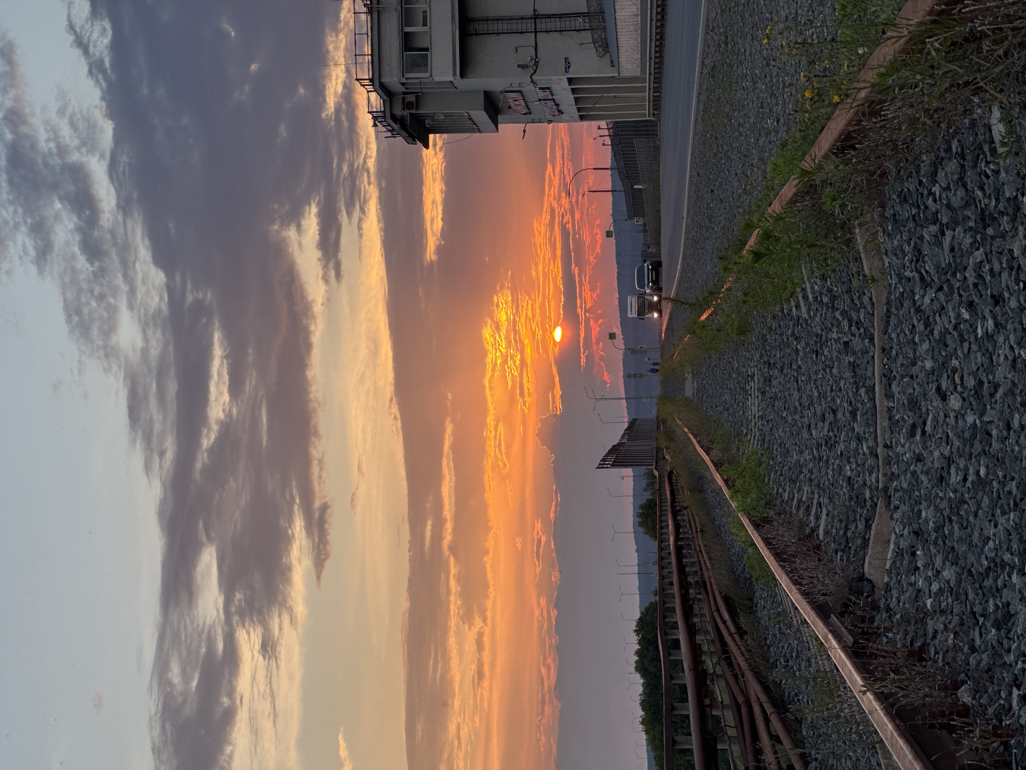 An outdoor vertical shot captures a railway track in the foreground, leading into the distance where a dramatic sunset paints the sky with vibrant orange and yellow hues. The sun is partially obscured by clouds, creating a glowing halo effect. To the right of the tracks, a multi-story building with a balcony railing and an attached structure with a sign is visible. Across the road, there are streetlights and some vehicles, including a truck and a car, driving away from the viewer. The scene appears to be a section of the Chaikovsky, Russia, railroad infrastructure during a sunset.