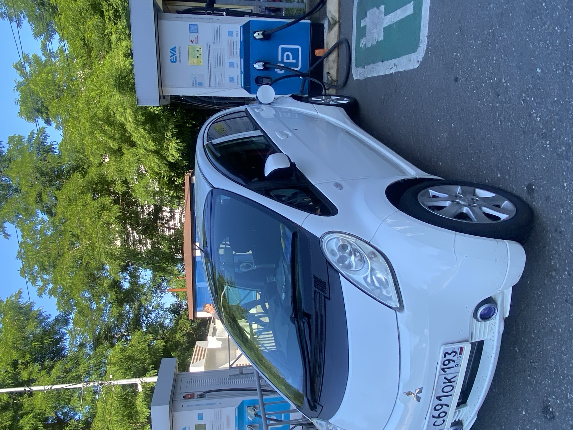 A white Mitsubishi i-MiEV electric vehicle is parked at a charging station in Sochi, Russia. The car is facing left and is plugged into a blue charging unit labeled "EVA intermobility". The charging station has a green symbol indicating it is an electric vehicle charging spot. In the background, lush green trees and a building are visible. The license plate of the car reads "C691OK 193 RUS". The scene appears to be outdoors on a sunny day.
