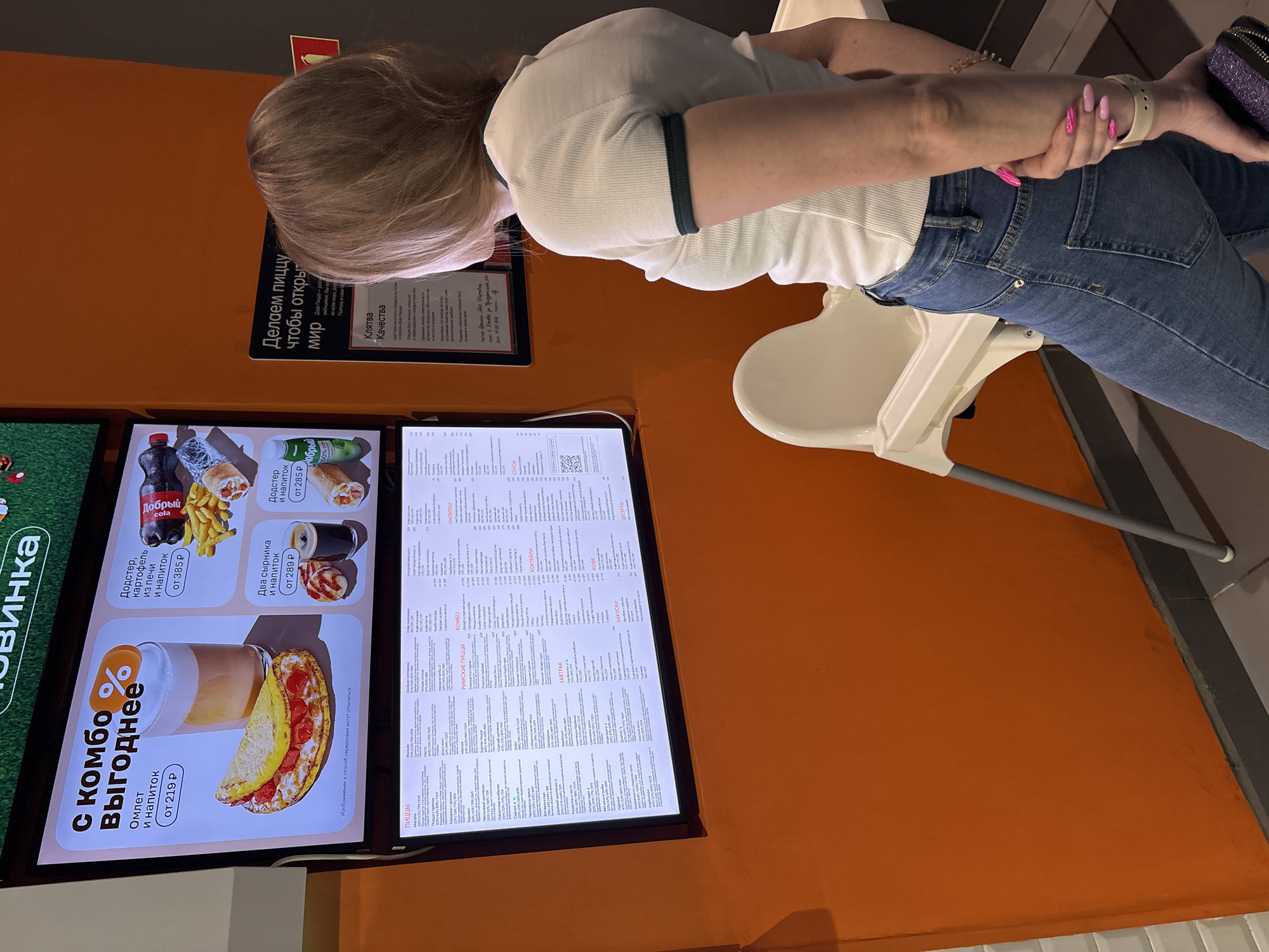 An indoor scene within a restaurant or cafe, identified as being in Izhevsk, Russia, is depicted. A single individual, viewed from behind, stands facing two vertically stacked illuminated digital menu boards. The person has light brown hair and is wearing a white, ribbed short-sleeved t-shirt with dark green trim on the collar and sleeves, and blue denim jeans. Their left hand crosses the body to grasp the right forearm, while the right hand holds a small, glittery purple object. The person's fingernails are long, pointed, and exhibit a pink gradient polish.
The upper digital display board shows promotional food combo deals with photographic images of items such as an omelet, a beverage, fried potatoes, and a wrap, accompanied by pricing in Russian Rubles. The lower digital display board presents a detailed menu, categorized with headings in Russian, including "ПИЦЦЫ" (Pizzas), "ЗАВТРАК" (Breakfast), "КОФЕ" (Coffee), and "ДЕСЕРТЫ" (Desserts).
To the right of the menu boards, a partially visible sign mounted on the orange wall displays Russian text, notably "Делаем пиццу чтобы открыть мир" (We make pizza to open the world) and "Клятва Качества" (Quality Oath). An unoccupied white plastic high chair with gray metal legs is positioned in the foreground, to the left of the person. The floor is covered with gray tiles. A small, rectangular red object is affixed to the wall on the far right of the frame. The dominant wall color is a solid orange.