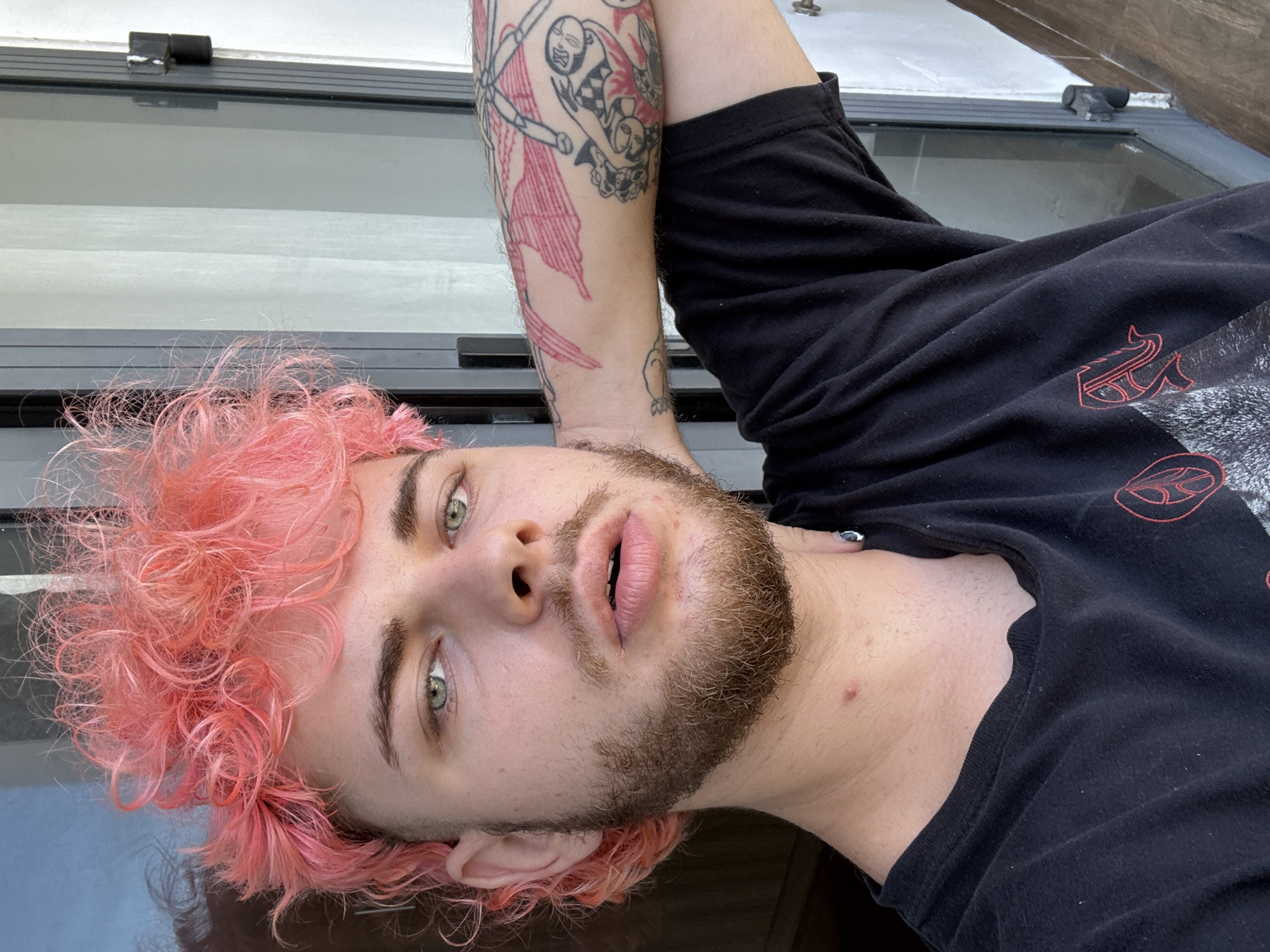 A close-up, low-angle shot of a person lying on a reflective surface, likely a table or counter. The person has pink curly hair, fair skin, a beard, and green eyes. They are wearing a black t-shirt with a red design on the right shoulder. The person's left arm is raised, revealing tattoos in red ink. The background includes a window and a white wall. There is no evidence of the location being in the city of Da Nang, Vietnam.