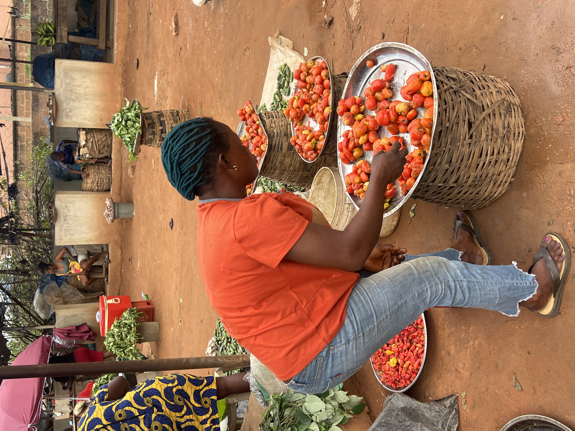The image depicts an outdoor market scene in Nkpor, Nigeria. A woman with blue dreadlocks, wearing an orange t-shirt and distressed blue jeans, is seated on the ground. She is actively engaged with a tray of red tomatoes, her hands reaching to sort or select them. Several large woven baskets are visible, some filled with produce like leafy greens and more tomatoes. In the background, other individuals are present, with one person tending to goods in a basket. Structures with corrugated metal roofing and concrete walls suggest a permanent or semi-permanent market stall. The ground appears to be made of packed dirt, and the lighting suggests it is daytime, with no strong indication of weather conditions beyond what might be typical for a warm, potentially humid climate. The overall impression is of a busy marketplace where local vendors sell fresh produce.