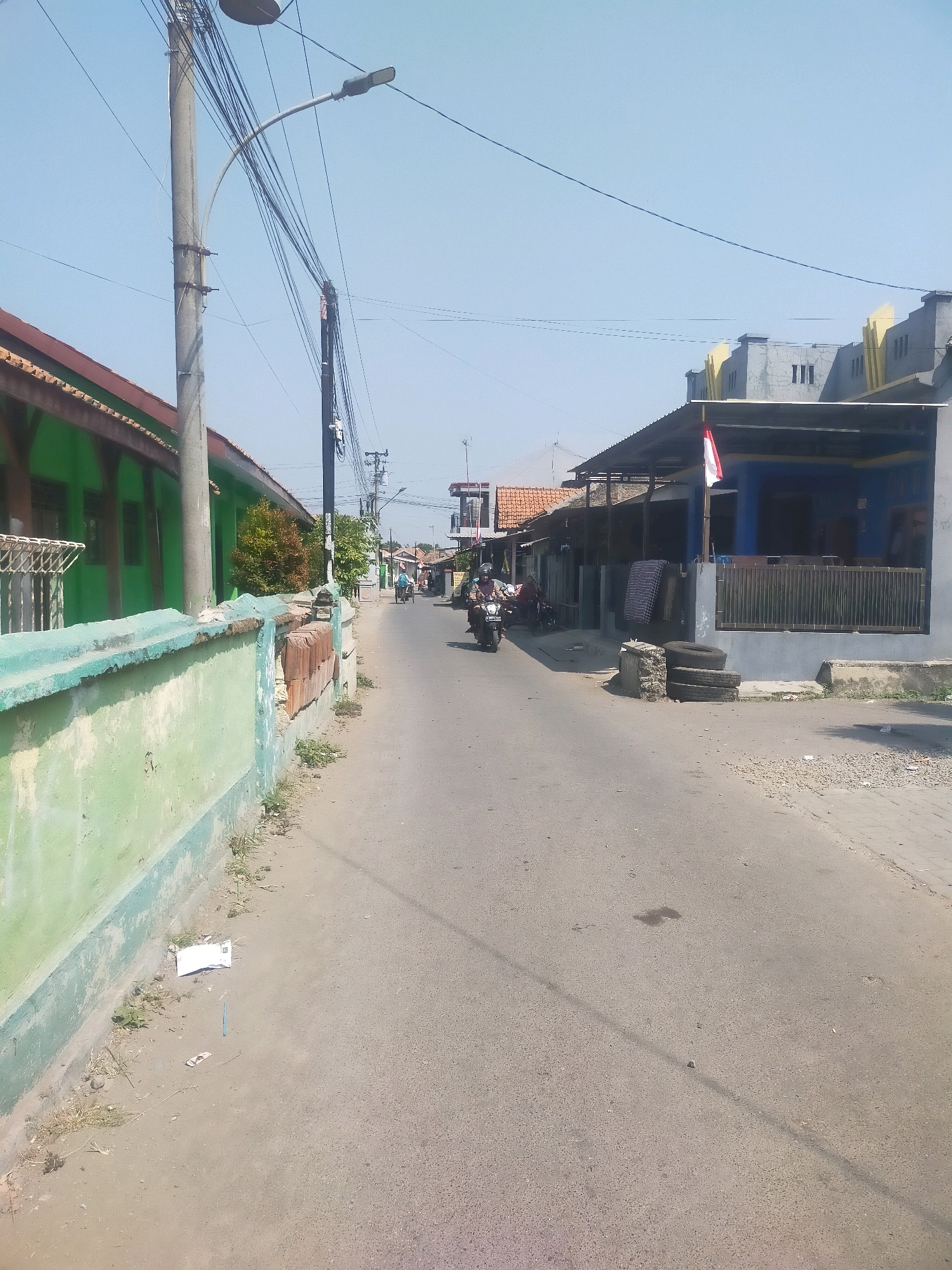 This image shows a street scene in Kabunan, Indonesia. The street is paved and appears to be in a residential or commercial area. On the left side of the street, there is a low green wall with a light blue trim, and behind it, a building with a green facade and a brown roof. Further down the street on the left, there is a light pole with multiple wires extending from it. On the right side of the street, there are several buildings with corrugated metal roofs. One building has a blue facade and a dark fence in front of it, with a small Indonesian flag displayed. A few people are visible on the street, including two individuals riding a motorcycle. One person is wearing a helmet. The sky is clear and blue. The overall impression is that of a typical street in a warm climate.