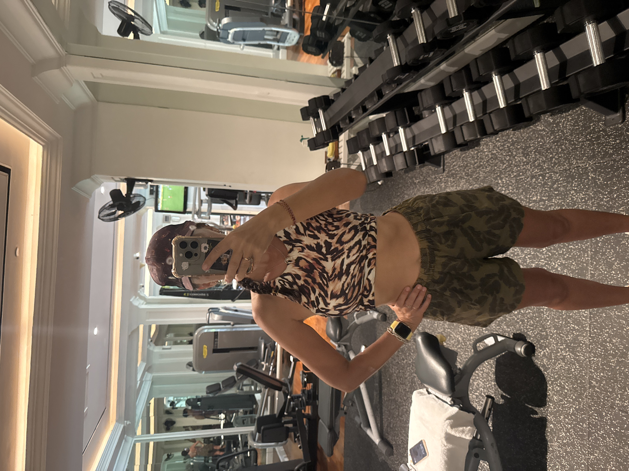 The image is a selfie taken in a gym. The subject is a woman in fitness apparel, wearing a printed top and camouflage shorts, holding a smartphone to capture her reflection in a large mirror. The gym is well-equipped with various exercise machines, including weight racks stocked with dumbbells. The background includes elliptical machines and treadmills. Based on the details of the architecture and gym equipment, there is no evidence to specifically determine the location within Kuta Selatan, Indonesia.