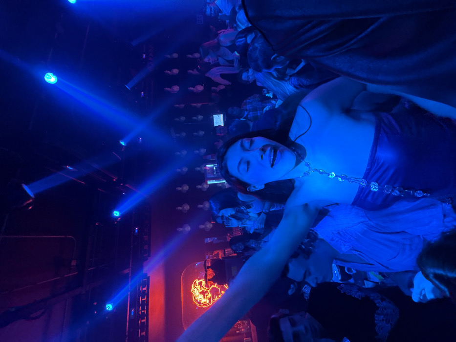 A woman in a purple dress and a crystal necklace is partying in a nightclub. Blue lights illuminate the scene, casting a vibrant glow. The crowd is dense around her. A sign, possibly for liquor, glows orange in the background. The woman seems to be enjoying the event, possibly singing or shouting in excitement. Other people can be seen around her, some wearing dark clothing.