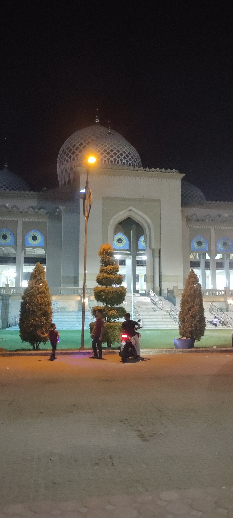 The image depicts a night scene in Lhokseumawe, Indonesia, featuring the Islamic Center. In the foreground is a paved road, in front of a strip of grass with trees and bushes. Three individuals are visible: a child to the left, an adult standing, and another adult on a motorbike. The background is dominated by the illuminated Islamic Center with its distinctive dome. A light pole stands between a tree and the building.