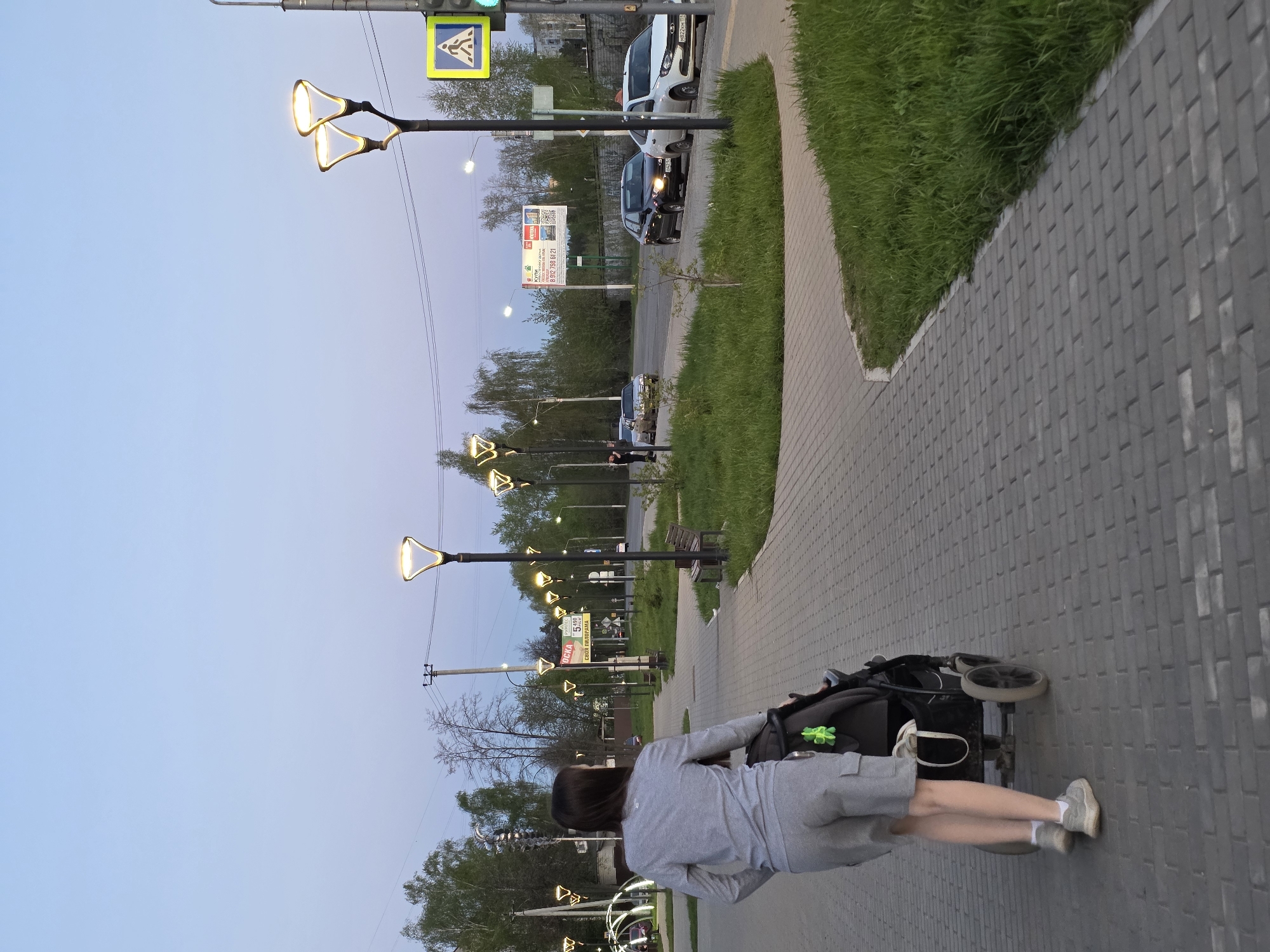 A medium shot captures a woman walking on a pedestrian sidewalk in Chaikovsky, Russia. She wears a grey ensemble and light sneakers, and is pushing a black baby stroller. The sidewalk is constructed of interlocking grey pavers, running adjacent to a patch of green grass. A row of modern streetlights illuminates the walkway. In the background, parked cars are visible along the street, and a sign displaying a large advertisement is noticeable. The image is oriented vertically, taken during twilight, evidenced by the cool blue tones in the sky.