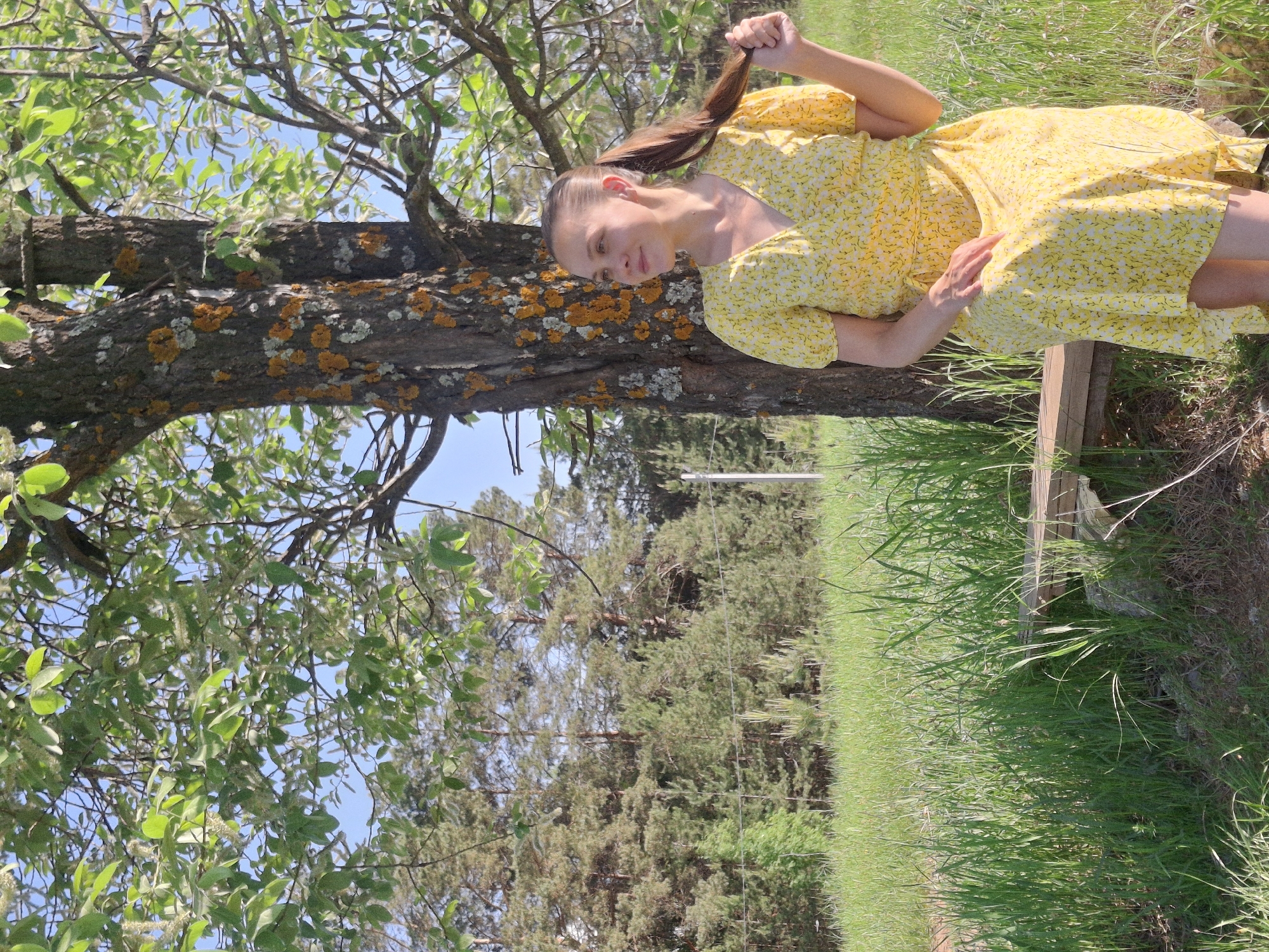 A woman wearing a yellow floral dress is posed by a tree in an outdoor setting. The tree trunk, covered in lichen, is prominent in the foreground, with leafy branches reaching upwards. In the background, a field of green grass stretches towards a line of trees. The location is a park or outdoor area in Chaikovsky, Russia.