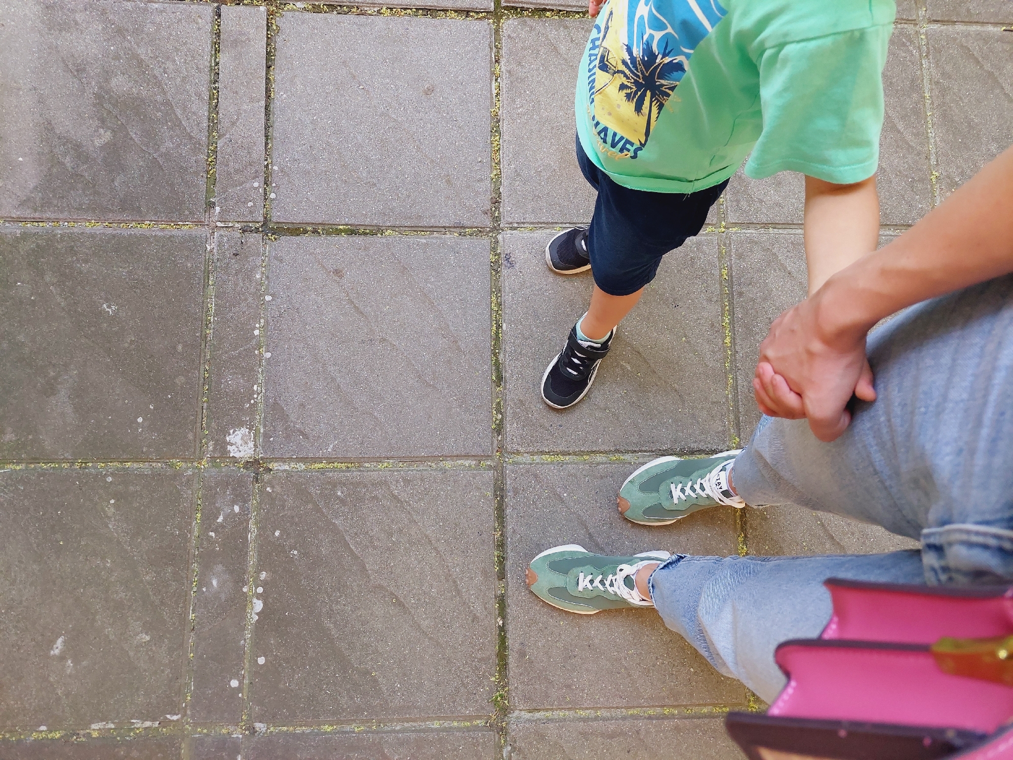 Two people, an adult and a child, are standing on a paved area made of square concrete tiles. The adult, on the left, is wearing light blue jeans and green sneakers. The child, on the right, is wearing dark blue shorts, black sneakers, and a light green t-shirt with a tropical print. The adult is holding the child's hand. The scene appears to be outdoors in an urban environment, possibly a park or a pedestrian area. The concrete tiles show signs of wear and tear, with some moss or dirt in the grout lines. The lighting suggests it is daytime. There is no information in the image to indicate the location is in Sofia, Bulgaria.