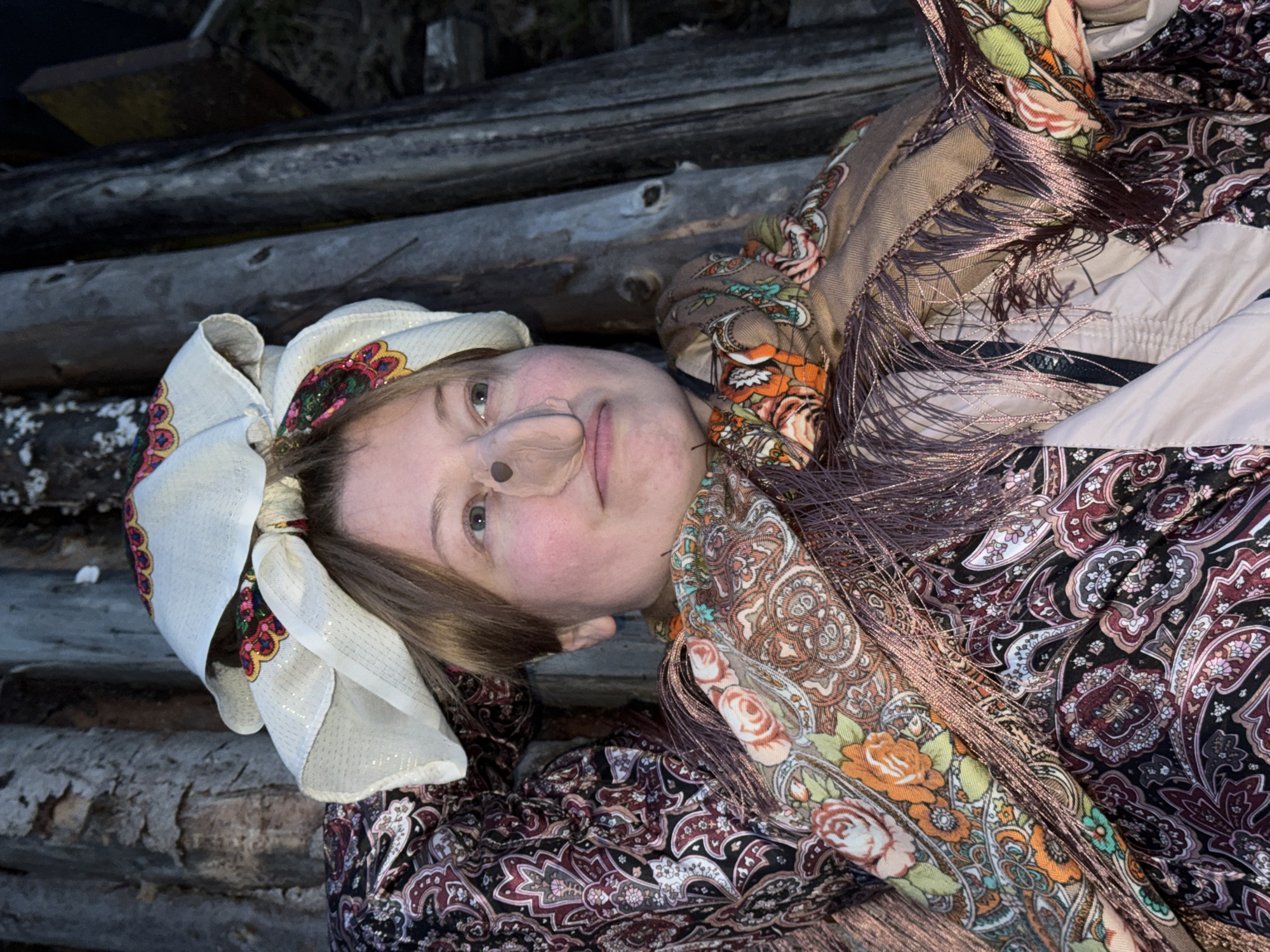 The image shows a person, likely a woman, lying on their back and looking up towards the camera. Her head is positioned towards the left side of the frame. She is wearing a white headscarf adorned with vibrant, colorful embroidery or print, featuring floral and stylized patterns. A light brown prosthetic resembling a pig's snout is placed on her nose. Her expression is calm, with a slight upturn at the corners of her mouth.
She is dressed in layers of patterned clothing. A prominent shawl, in shades of brown with intricate floral and paisley designs in orange, green, and pink, is draped over her. This shawl has numerous shimmering, dark brown fringes along its edges. Beneath the shawl, another garment with a dark background and white, purple, and reddish-brown paisley patterns is visible.
The background consists of several dark, weathered wooden logs, suggesting an outdoor, rustic environment, possibly a rural setting or near a wooden structure. The lighting appears dim, indicative of twilight or evening conditions. No text is visible in the image. This scene is set in Вассята, Russia.