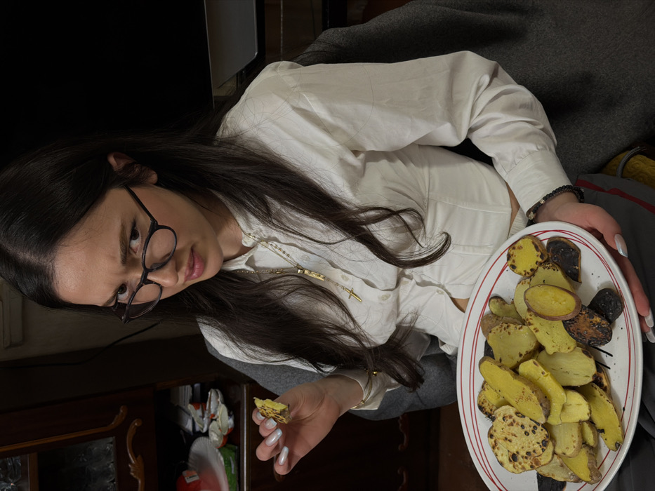 An indoor scene in Arinj, Armenia, depicts a young adult female seated, holding a plate of cooked food. The subject has long, dark hair, wears eyeglasses with dark frames, and is dressed in a white button-up shirt and dark trousers. A gold chain with a cross pendant and a rectangular pendant is visible around her neck, along with a dark beaded bracelet on her left wrist. Her fingernails are manicured with a light polish. She holds an oval white plate containing numerous slices of cooked root vegetables, primarily yellow and purple with some charred portions, in her left hand. In her right hand, she holds a single piece of similar cooked vegetable. Her gaze is directed towards the viewer with a furrowed brow and a questioning or skeptical expression. The background on the left features a dark wooden cabinet with glass doors, displaying indistinct contents, and a surface below holding white plastic objects, possibly opened confectionery containers. The background on the right is dark and indistinct, with a portion of a grey fabric object visible behind the subject.