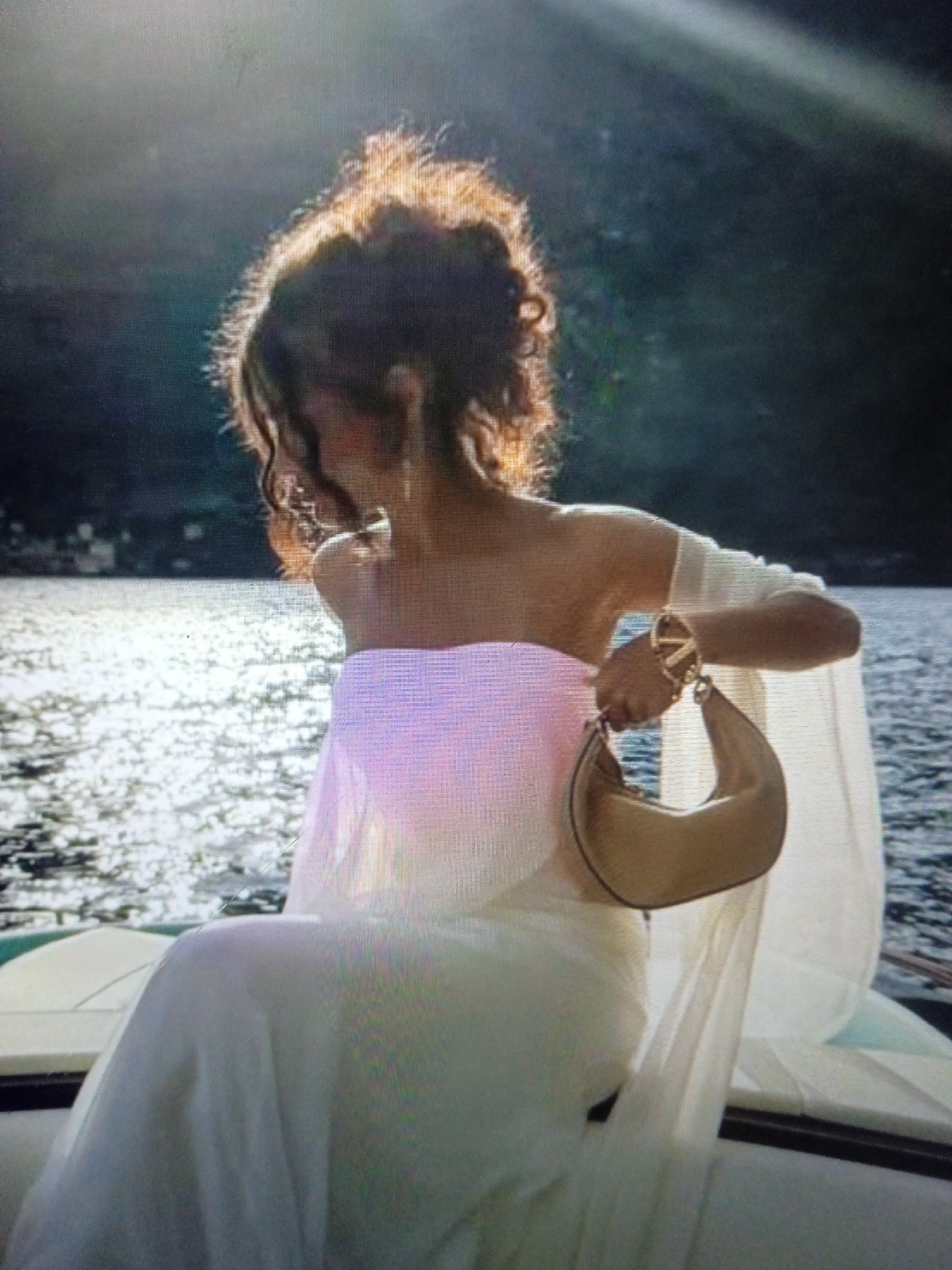 The image shows a person from behind, wearing an off-the-shoulder white dress and holding a beige crescent-shaped handbag with a gold chain strap. The person's hair is dark and styled in an updo. They are seated on what appears to be a boat, with shimmering water in the background and a dark, mountainous landscape further beyond. The sunlight is strong, casting a glow on the water and the person's hair. There is no visible indication of the location being Chernivtsi, Ukraine, or any urban environment.
