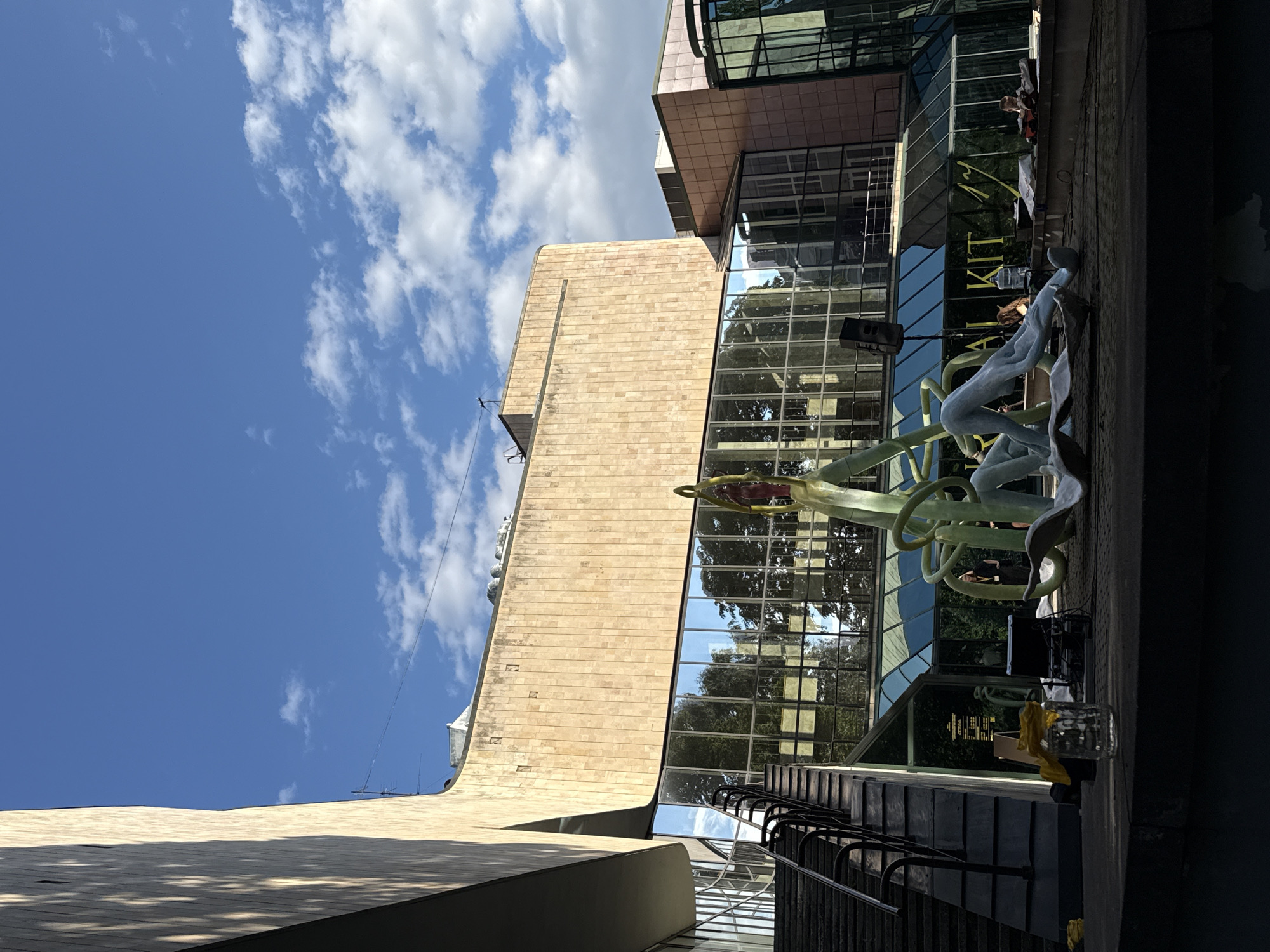 The image shows a building with a modern architectural style, characterized by large glass windows and beige stone cladding. In the foreground, there is a large abstract sculpture, seemingly made of light blue and green material, with a red figure at its top. Various objects, including a speaker, a water bottle, and what appears to be a glass jar with yellow fabric, are scattered on the ground in front of the sculpture and the building. A set of dark metal stairs is visible on the left side of the image, leading up towards the building. The sky above is blue with scattered white clouds. The building's facade reflects trees and the sky. People are sparsely visible in the background, near the building. The scene appears to be set in an outdoor urban environment. This is the National Library of Latvia in Riga, Latvia.
