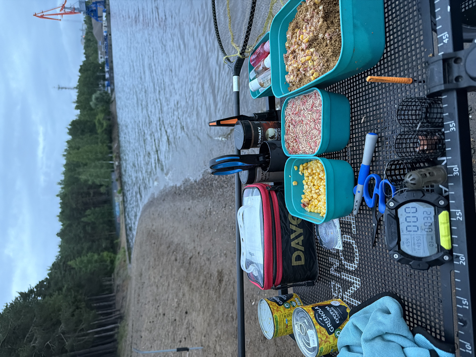 This image presents a detailed fishing setup positioned on a black perforated metal fishing station table, overlooking a sandy beach and a body of water in Chaikovsky, Russia. The sky is overcast, providing diffuse lighting.
In the immediate foreground, various fishing provisions and tools are arranged on the black table, which features numerical markings from 10 to 45 along its bottom edge. Two yellow cans of "GREENOLA" sweet corn, with Cyrillic text "КУКУРУЗА САХАРНАЯ" (Sweet Corn), are visible, one of which is open. A black and red cooler bag, branded "DAVE", with a clear lid, is situated next to the cans. Adjacent to this, three teal-colored plastic containers hold bait: one with yellow corn kernels, another with a mixture of pink and red maggots, and a third with a brown granular groundbait mixture containing corn.
Additional items on the table include a pair of blue-handled scissors, a black container holding more tools (including another pair of blue-handled scissors and a black-and-orange fishing tool), and a black jar labeled "DUNAEV" and "ORANGE POWDER". A spooled fishing line is visible. A crumpled blue microfiber towel lies on the left. A black "MATRIX" branded digital timer/scale, with yellow accents, displays "TIME: 4:54", "ET/COUNT: 143.53", "NET: 000", "03", and "0032". Three black fishing feeders/cages are placed near the timer, along with a bright orange plastic baiting tool. Behind the main bait containers, small clear and colored plastic bottles, likely containing liquid attractants, are visible next to a black fishing net.
The midground features a sandy beach leading to the body of water, which exhibits slight ripples on its surface. A metal fishing rod holder is partially visible, inserted into the sand on the left side of the frame.
The background shows a dense forest of tall pine trees lining the left side of the shoreline. Further along the right side of the water, an industrial area is apparent, marked by a prominent red and blue crane and a metal communications tower, suggesting proximity to a port or industrial facility within Chaikovsky. No individuals are visible in the scene.