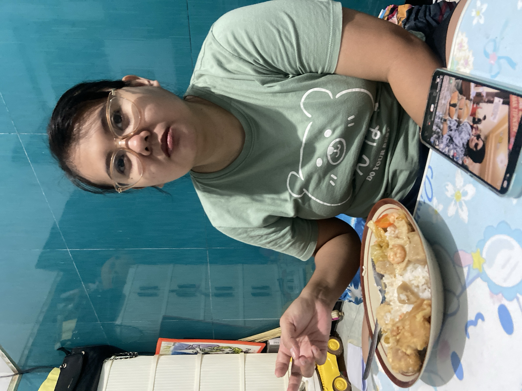 A medium shot shows a person with brown hair, wearing glasses and a green T-shirt with a bear design, sitting at a table and looking at the camera. On the table is a bowl of food, a cell phone displaying a video, and other miscellaneous items. The background features a light blue wall. The location is Gelam, Indonesia.