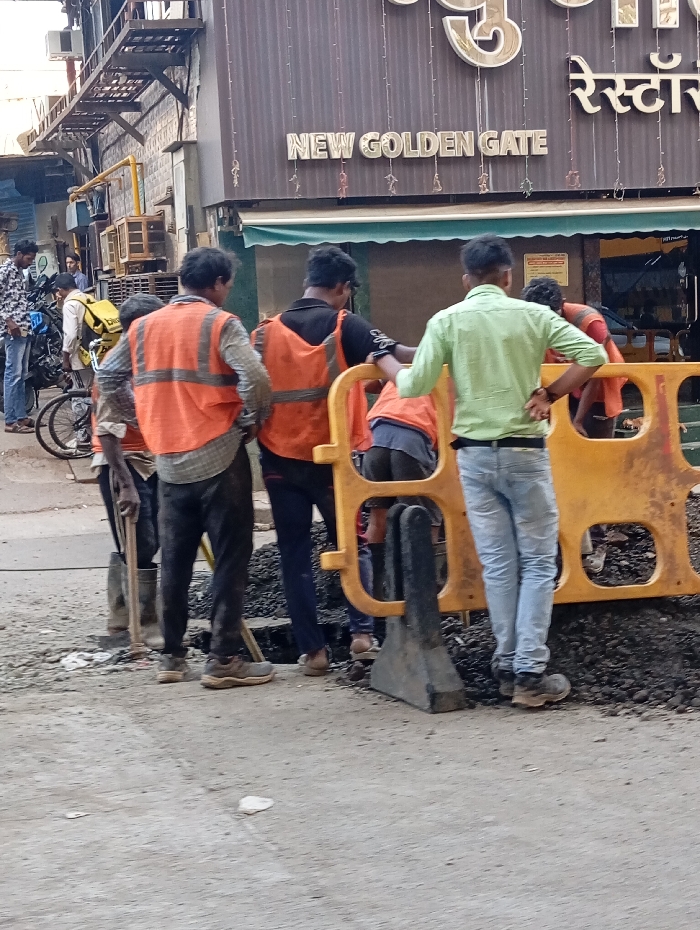 The scene depicts an urban street in Mumbai, India, in front of a building identified by a prominent sign as "NEW GOLDEN GATE RESTAURANT." In the foreground, four adult males are gathered around an excavated area on the asphalt road, marked by a yellow plastic road barrier with black bases. Three of these individuals wear orange high-visibility safety vests over their attire. One man in an orange vest is positioned with a long-handled tool, possibly a shovel, near a mound of dark, loose material consistent with excavated road debris. Another man in an orange vest is seen bending over the excavated section. A third man, wearing a light green collared shirt and blue jeans, stands upright with his hands on his hips, observing the activity. In the background on the left, a man in a patterned shirt stands next to a blue bicycle equipped with a large yellow delivery bag. Additional individuals and motorcycles are partially visible further down the street. The restaurant building features a brown corrugated metal facade and a green awning. The environment suggests an active urban street with ongoing roadwork.
