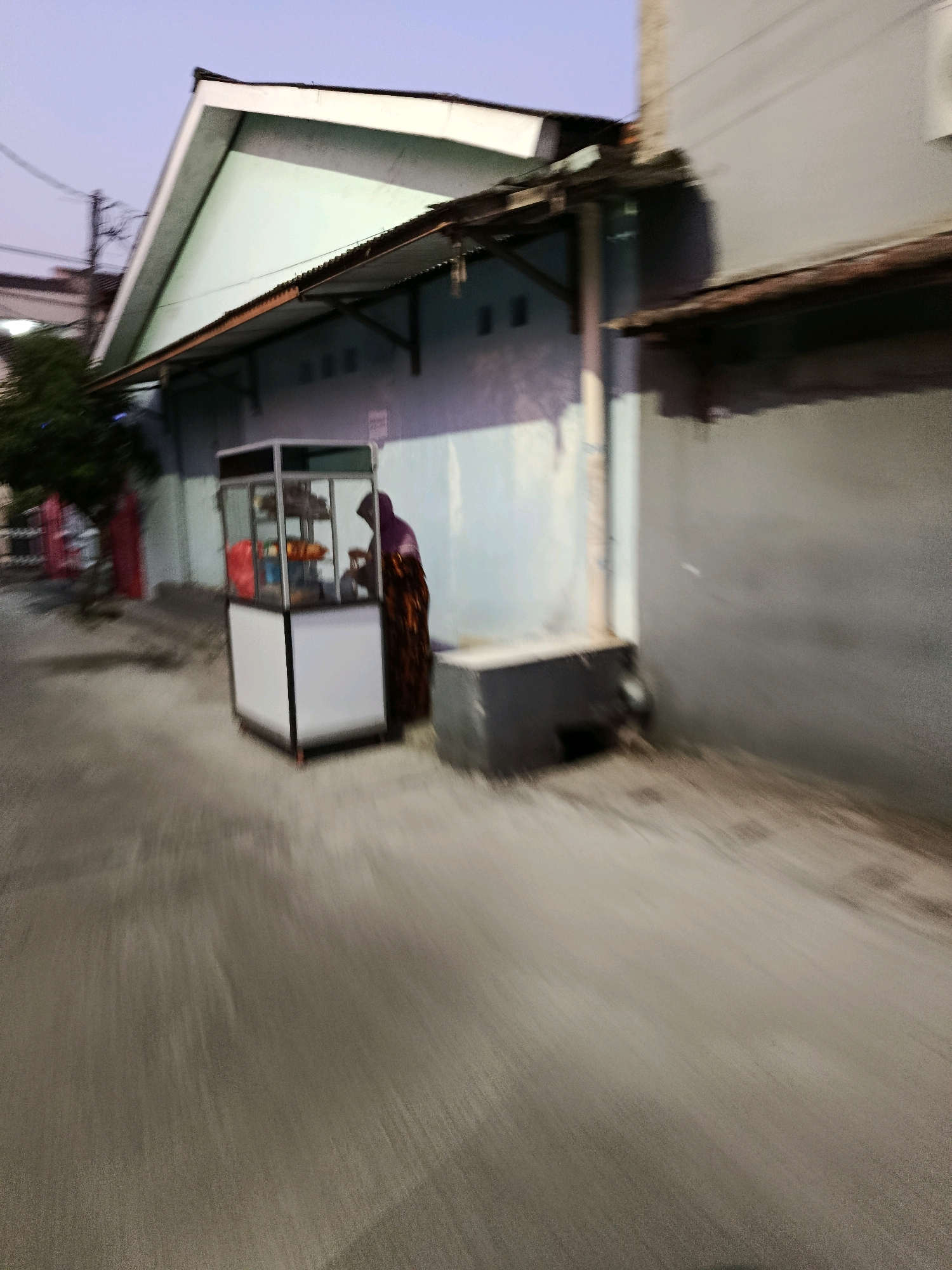 The image depicts an outdoor scene in the city of Bekasi, Indonesia, likely on a street or alleyway. A food cart, appearing to be made of metal and glass, is visible in the center of the frame. A person, wearing a headscarf and a patterned dress, stands behind the cart. The cart is stocked with various items, possibly food or beverages, though their exact nature is unclear due to the image's blurriness.
To the left of the cart, a portion of a building with a green roof and a light blue wall is visible, along with a tree. To the right of the cart, a grey building with a dark roof overhang occupies a significant portion of the frame. The ground appears to be asphalt or a similar paved surface. The overall image is characterized by motion blur, suggesting it was taken while moving, possibly from a vehicle. The lighting indicates it is either early morning or late evening.