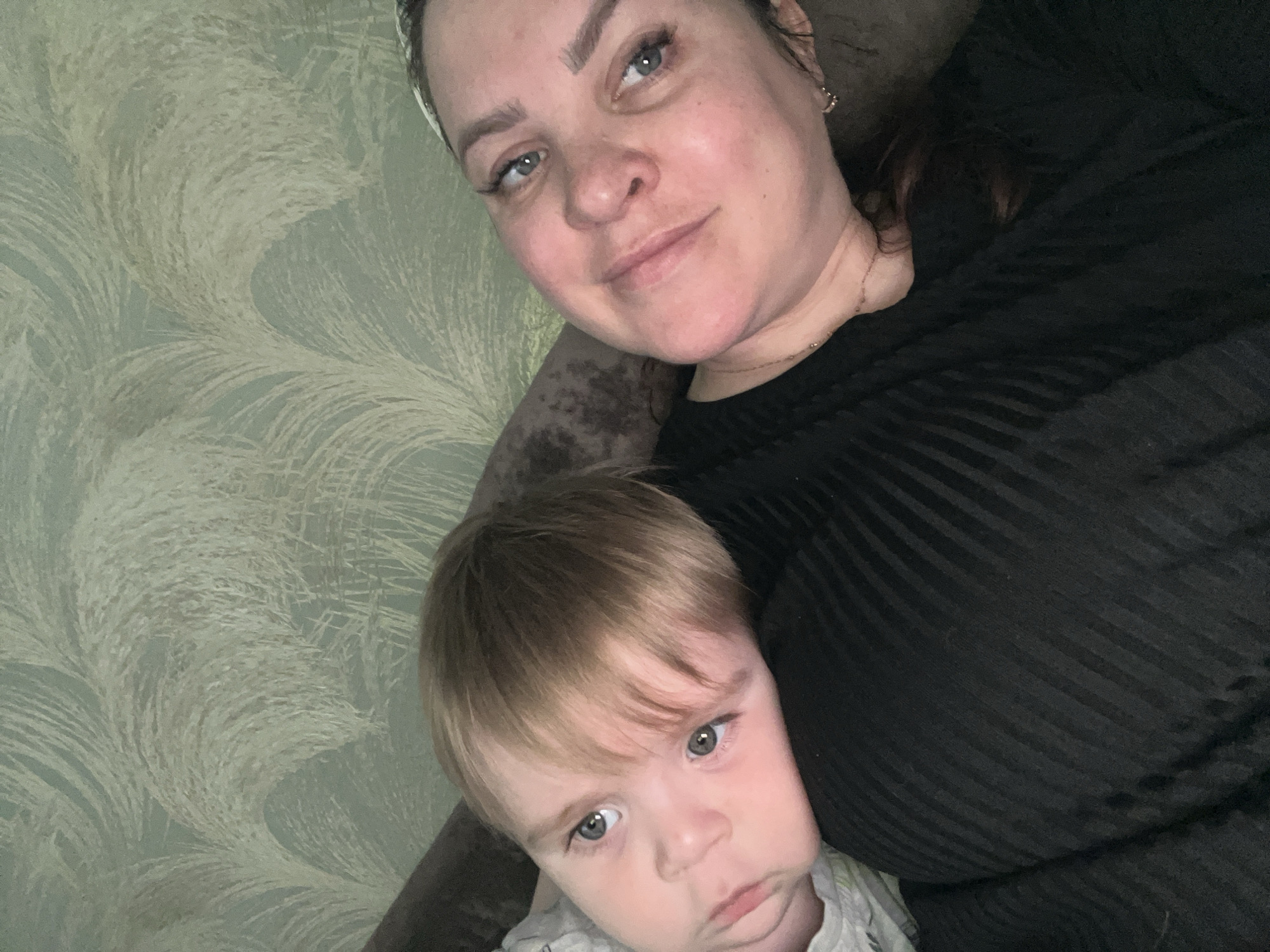 The image depicts an adult female and a young male child, likely a mother and son, in an indoor domestic setting. The scene is captured from a slightly elevated, oblique angle, typical of a selfie or close-up photograph.
The female, positioned on the right, has fair skin, light blue-grey eyes, and dark eyebrows. Her dark hair is partially visible and appears to be held back by a light-colored headband or scarf. She is wearing a black top with vertical ribbed or striped texture and a delicate gold-toned necklace. Her expression is a subtle, closed-mouth smile, and she is looking directly at the camera.
The child, positioned on the left and leaning against the female, has fair skin, light blonde hair with bangs, and light blue-grey eyes similar to the female. He is wearing a light-colored garment, possibly white or light grey, with a faint, indiscernible pattern. His head rests on the female's shoulder or chest, and his expression is neutral or slightly pensive, with his gaze directed towards the camera.
The background consists of a wall covered in wallpaper featuring a light blue-green base color with a recurring pattern of swirling, feather-like, or abstract organic shapes in off-white or light beige. A dark, upholstered piece of furniture, possibly a sofa or armchair, is visible in the lower left and right corners, indicating where the individuals are reclining. The lighting is soft and ambient.