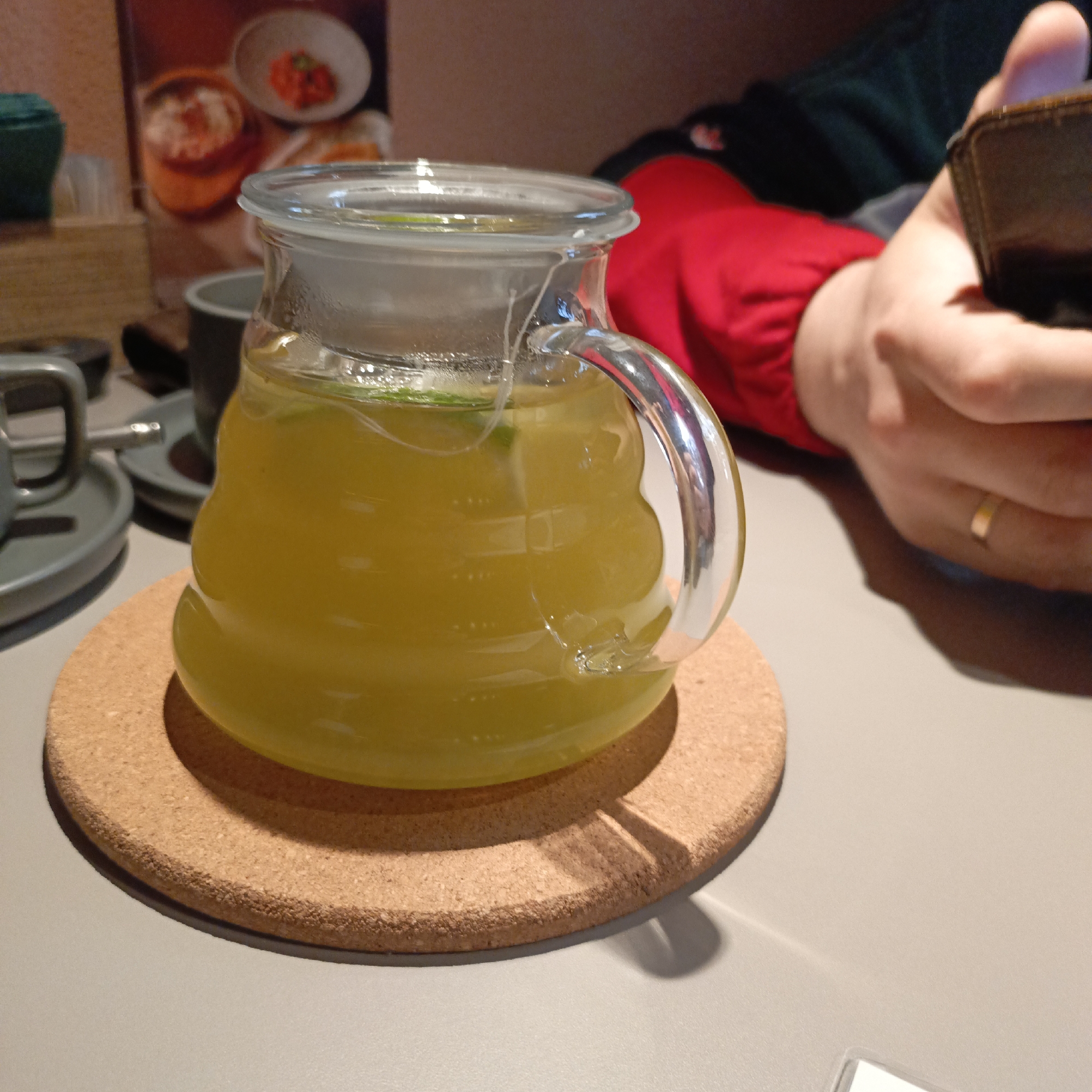 The image presents an indoor scene, likely a cafe or restaurant, centered around a clear glass pitcher filled with a bright yellow beverage, possibly a fruit tea or lemonade. The pitcher has a distinctive ribbed design on its lower body and a clear handle, and it sits on a round, thick cork coaster with a dark rim. Small green specks, likely mint or lime, are visible within the yellow liquid, and the upper part of the pitcher shows slight condensation, suggesting a cold drink.
In the blurred background to the right, a person's hand is visible, holding a brown wallet or phone, and a gold ring adorns one of their fingers. This person is wearing a red long-sleeved garment and a dark top. On the left side of the table, other dark-colored ceramic mugs or cups and saucers are partially visible. A warm brown wall in the far background displays a blurred poster or menu with images of food dishes in bowls. The light grey tabletop reflects subtle shadows from the pitcher and other items. The scene depicts a relaxed moment, possibly during a meal or break. No legible text is visible.