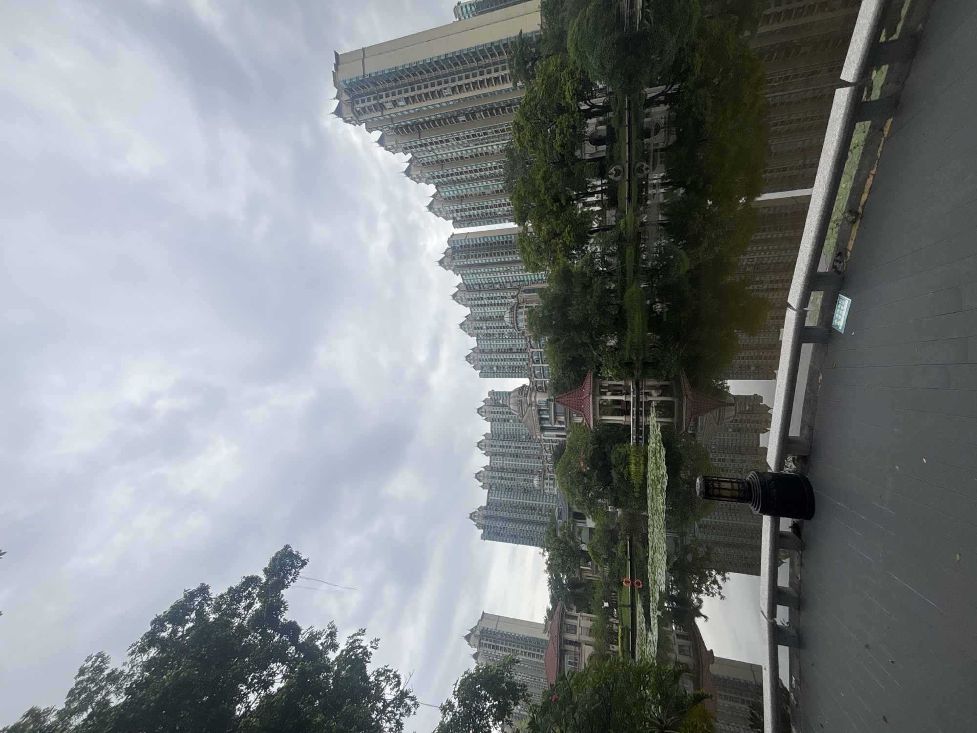 The image depicts a serene waterscape in the city of Nanhai District, China, likely within a residential complex or park. In the foreground, a dark grey paved walkway curves to the right, bordered by a low, grey stone wall with decorative carvings. A black lamp post stands on the walkway near the wall. Beyond the walkway, a calm body of water reflects the surrounding environment and the sky. Lily pads dot the surface of the water in the midground.
In the midground, a small, ornate pavilion with a red, hexagonal roof and white columns stands beside the water. It is surrounded by lush green trees and vegetation. In the background, several tall, modern apartment buildings rise against a cloudy sky. These buildings are characterized by their numerous floors, similar architectural styles, and balconies. Some older, more traditional buildings with red roofs are also visible amongst the trees, adding a contrast to the modern high-rises. The overall impression is one of urban development integrated with natural and recreational spaces. No people are visible in the image.