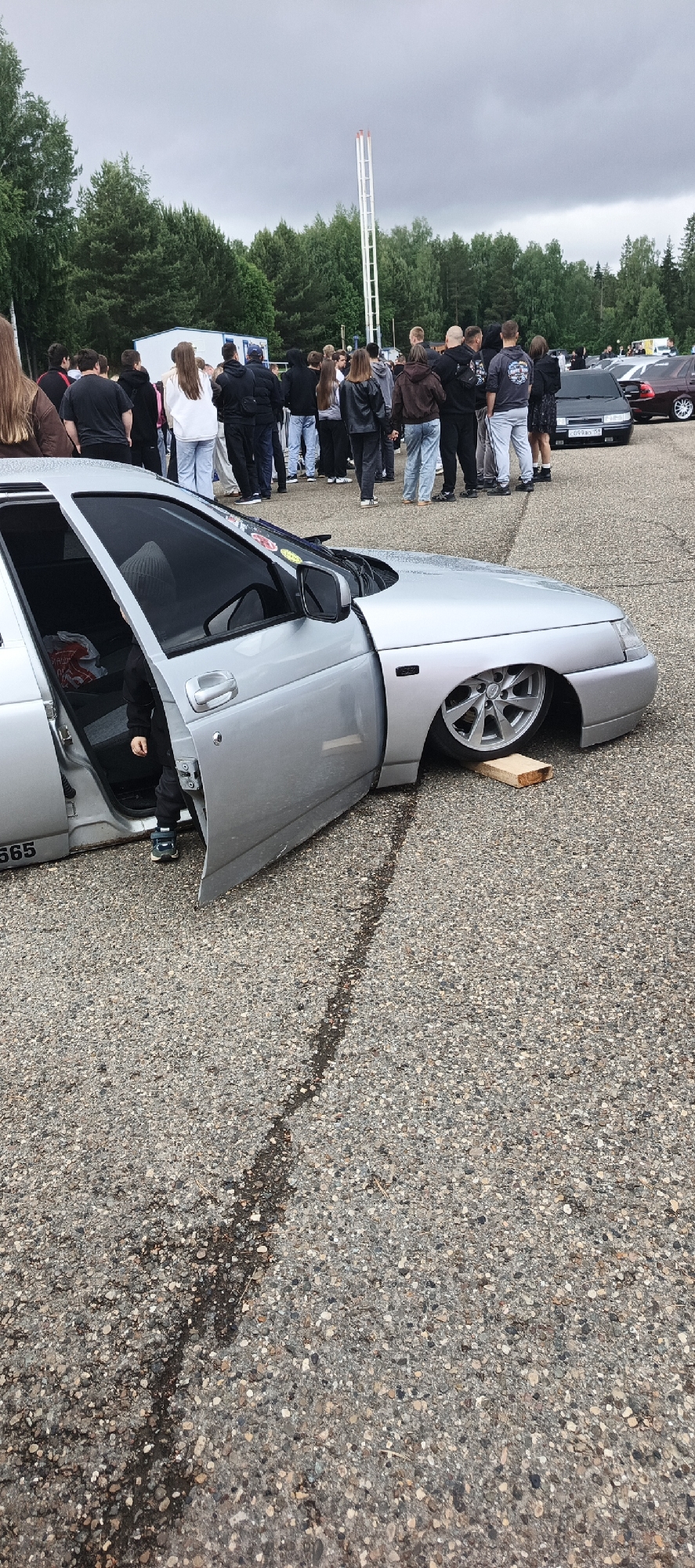 The image depicts an outdoor car event or gathering in an unknown location, characterized by a paved surface, a surrounding forest, and an overcast sky.
In the foreground, a heavily modified, silver four-door sedan (likely a Lada 2110-series or similar vehicle common in Eastern Europe) is parked with its front left wheel extremely cambered and resting on a wooden block, indicating an "air ride" suspension system in its lowest position. The front left door is wide open, revealing a small child inside, wearing dark clothing and blue and green sneakers, standing or sitting low within the car. A white plastic bag with red lettering is visible on the passenger seat. The number "565" is printed on the lower rear portion of the car's body. The ground is a dark, coarse aggregate pavement (asphalt/gravel mix) with a distinct dark, slightly wet-looking crack or line running diagonally from the lower left to the mid-right.
In the midground, a large group of approximately 30-40 people, predominantly young adults, are gathered. Most are casually dressed in hoodies, t-shirts, jeans, and sneakers, standing and observing the parked cars or socializing. Their gazes are generally directed towards the cars or at each other. Behind this crowd, several other vehicles are visible, including a black sedan with a Russian-style license plate (C099AX 93) and a dark maroon or brown sedan with chrome wheels, both appearing to be similarly lowered or modified.
The background consists of a dense line of various trees, creating a forest-like border along the horizon. The sky is uniformly overcast and light gray, suggesting a cloudy day without direct sunlight. A tall, white, latticed metal tower with a red top structure stands prominently on the right side of the background, extending high above the treeline. A small, white, rectangular building or portable cabin is visible amidst the trees on the left side of the background.
The overall scene suggests a casual car enthusiast meet-up.
