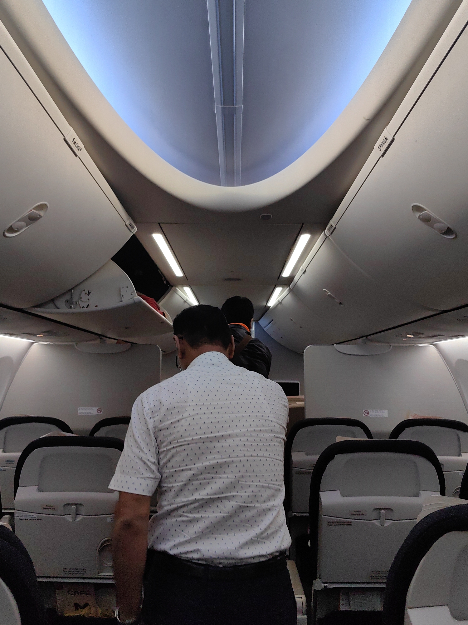 The image depicts the interior of an airplane cabin, viewed from behind a passenger. The passenger in the foreground is a male, wearing a white short-sleeved shirt with a pattern of small blue dots and dark trousers. He is walking down the aisle. Behind him, another passenger, also appearing to be male, is partially visible, wearing a dark jacket with an orange collar. The cabin features overhead luggage bins, with one bin open revealing some red-colored items inside. Fluorescent lights run along the ceiling, illuminating the aisle. Rows of airplane seats are visible on either side. The image does not provide any specific visual cues that would allow for the determination of its location within the city of Mumbai, India.