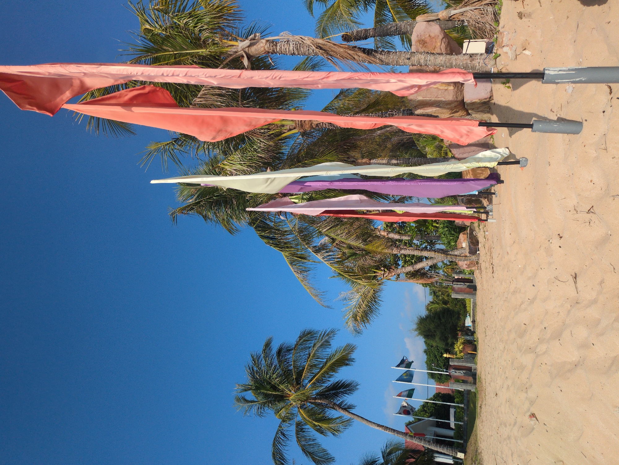 The image shows a sunny beach scene in Phú Quốc, Vietnam. The focus is on a row of colorful, triangular fabric flags attached to poles set in the sand. The flags appear in shades of pink, peach, light green, and purple. Behind the flags are palm trees with green fronds against a clear blue sky. In the distance, smaller flagpoles with national flags can be seen in front of a building. The foreground is sandy beach with visible footprints.