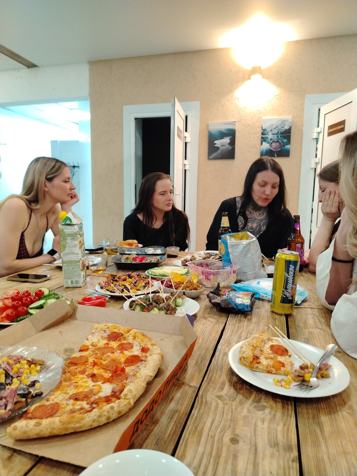 The image shows a group of five women sitting around a wooden table laden with food and drinks. The table is covered with a variety of items, including a half-eaten pizza in a cardboard box, a plate with a single slice of pizza and a side of corn, skewers, fresh vegetables, sushi, and a container of salad. There are also juice, beer bottles, a can of beer, and bags of chips on the table.
The women appear to be engaged in conversation or contemplation. The woman on the left is leaning on her hand, looking towards the center of the table. The woman next to her is looking slightly downward. The woman with the tattoo on her chest is looking down, perhaps at the food or her phone. The woman next to her is partially obscured, and the last woman is mostly out of frame.
The background features a light-colored wall adorned with three landscape paintings. There are two doorways on the wall. Based on the label on the beer can and the overall aesthetic, the location is likely in Chaikovsky, Russia.